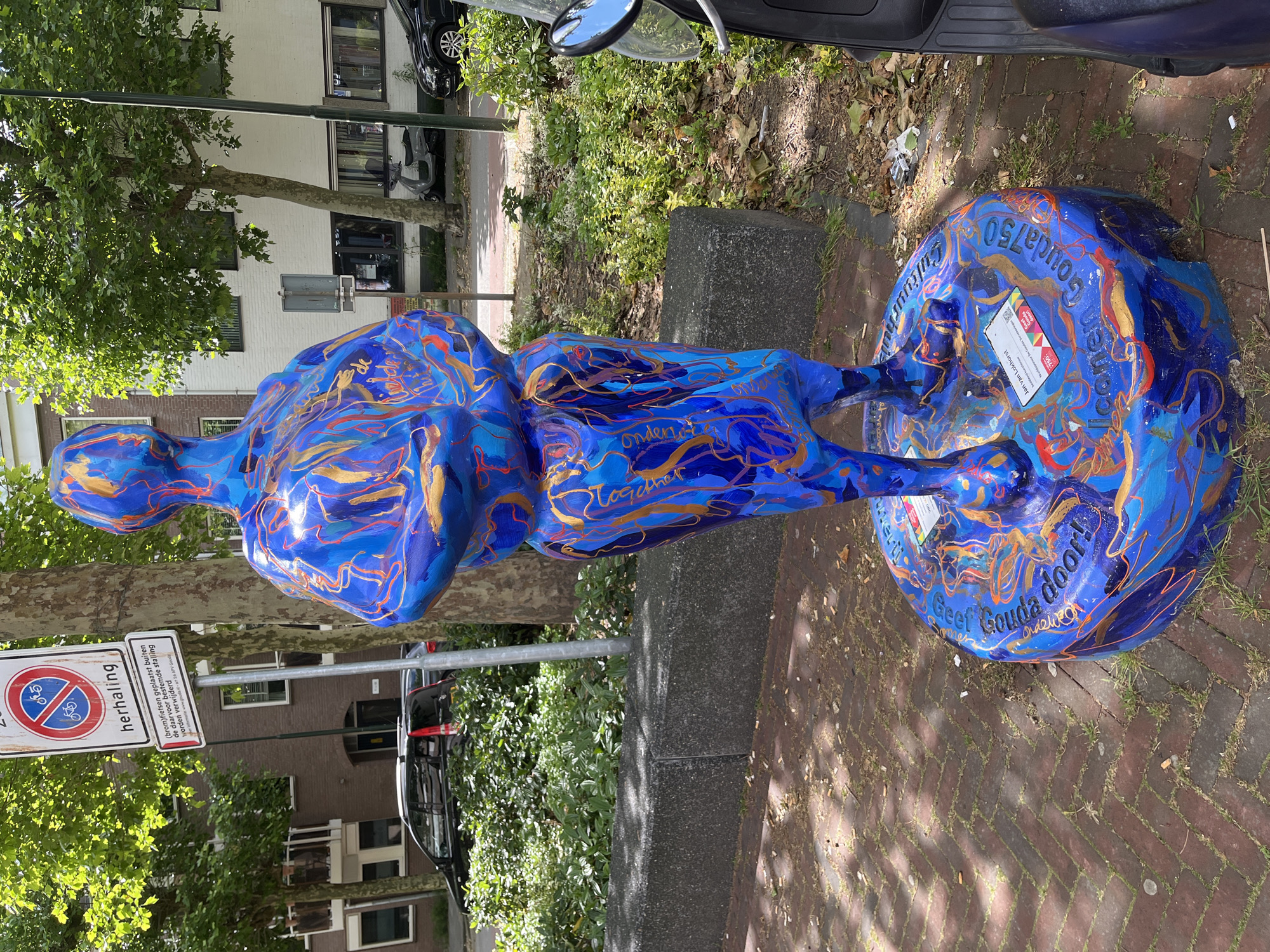 A vibrantly painted blue statue of a person with swirling gold and orange patterns stands on a circular base on a brick-patterned sidewalk. The statue is located in an urban environment with trees and buildings visible in the background. A street sign indicating "herhaling" with a bicycle and a motorcycle symbol crossed out is attached to a pole to the left of the statue. Text is visible on the base of the statue and on a small label attached to it. In the background, a black car, a scooter, and a dark-colored vehicle are parked. Green foliage and trees line the sidewalk and are visible behind a grey concrete planter. The ground is a reddish-brown brick pattern with some grass and scattered debris, possibly cigarette butts.
