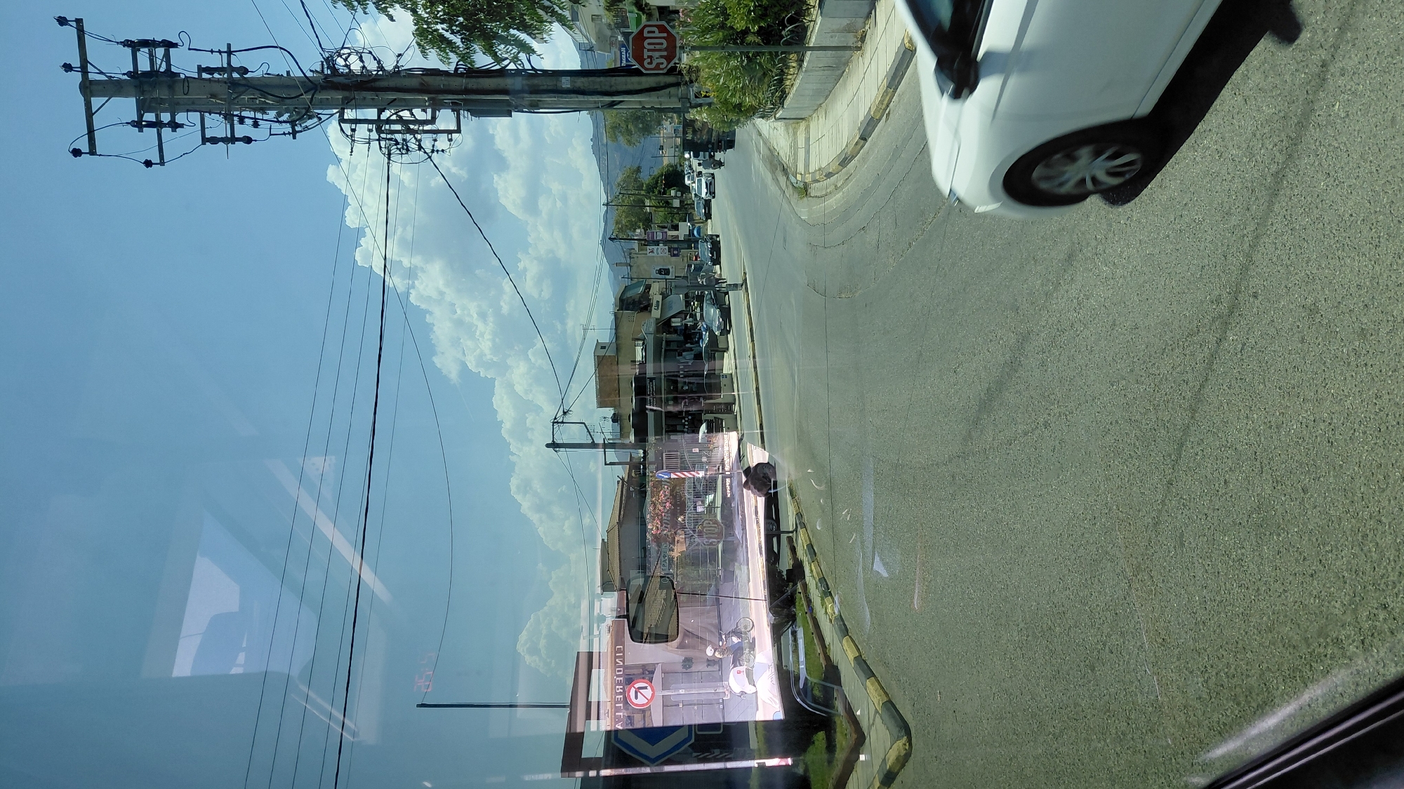 The image depicts a street scene in the Municipal Unit of Aegio, Greece, as viewed through a reflective surface, likely a window. The foreground shows a paved road with yellow and black striped curbs on the left. A white car is visible on the right side of the road, partially in frame.
In the midground, a street curves to the right. Several buildings line the street, appearing to house shops. Signs with Greek text are visible on the buildings. A traffic light or sign is visible on a pole on the right, and a red circular STOP sign is also present. Electrical poles and wires are prominent throughout the scene, crisscrossing the sky.
The background reveals hills or mountains under a bright blue sky with scattered white clouds. Reflections are visible on the surface through which the image is captured, showing what appears to be the interior of a vehicle, including a window frame and a hint of seating. One prominent reflection on the left side shows a storefront with a barber pole and a scooter with a rider in front of it. Another reflection displays text that appears to be "CINDERELLA" reversed.
There are no individuals clearly identifiable in the scene outside of the reflection of the scooter rider. The environment suggests a commercial area within a town or city, with clear daytime lighting.