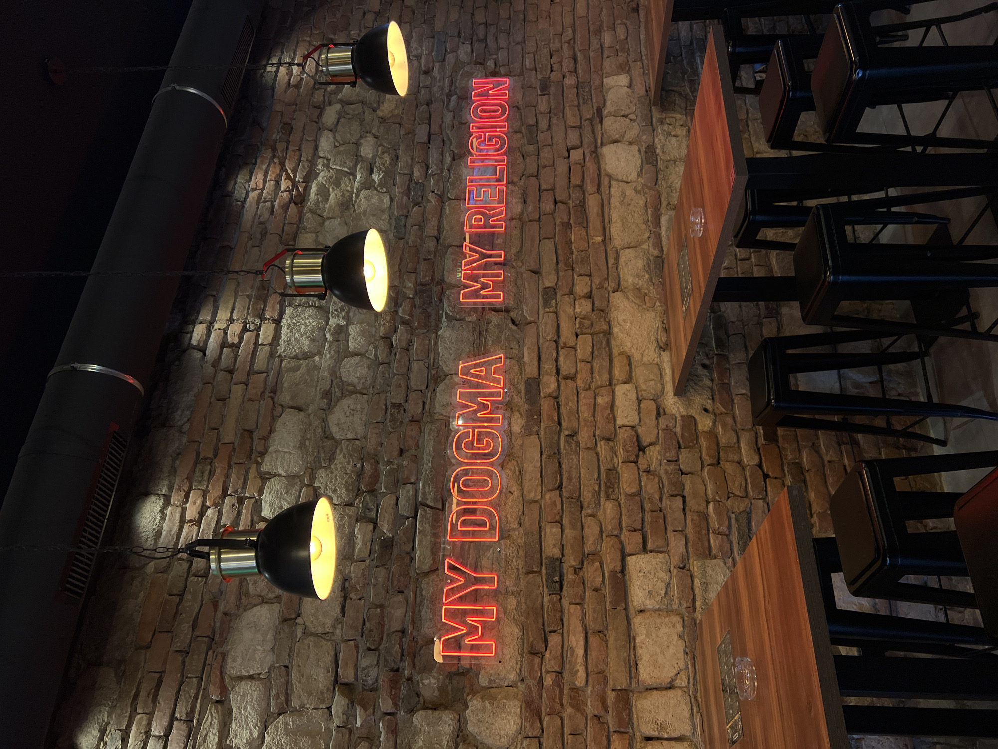 The image shows a dimly lit interior space, likely a bar or restaurant, with a rough brick wall as the primary backdrop. Three industrial-style lamps are mounted on the wall, each casting a warm glow. Between the lamps are two vertically stacked neon signs, reading "MY DOGMA" and "MY RELIGION." To the right of the wall are wooden tables with black metal legs and black chairs. The location is the city of Belgrade, Serbia. No people are visible in the image.
