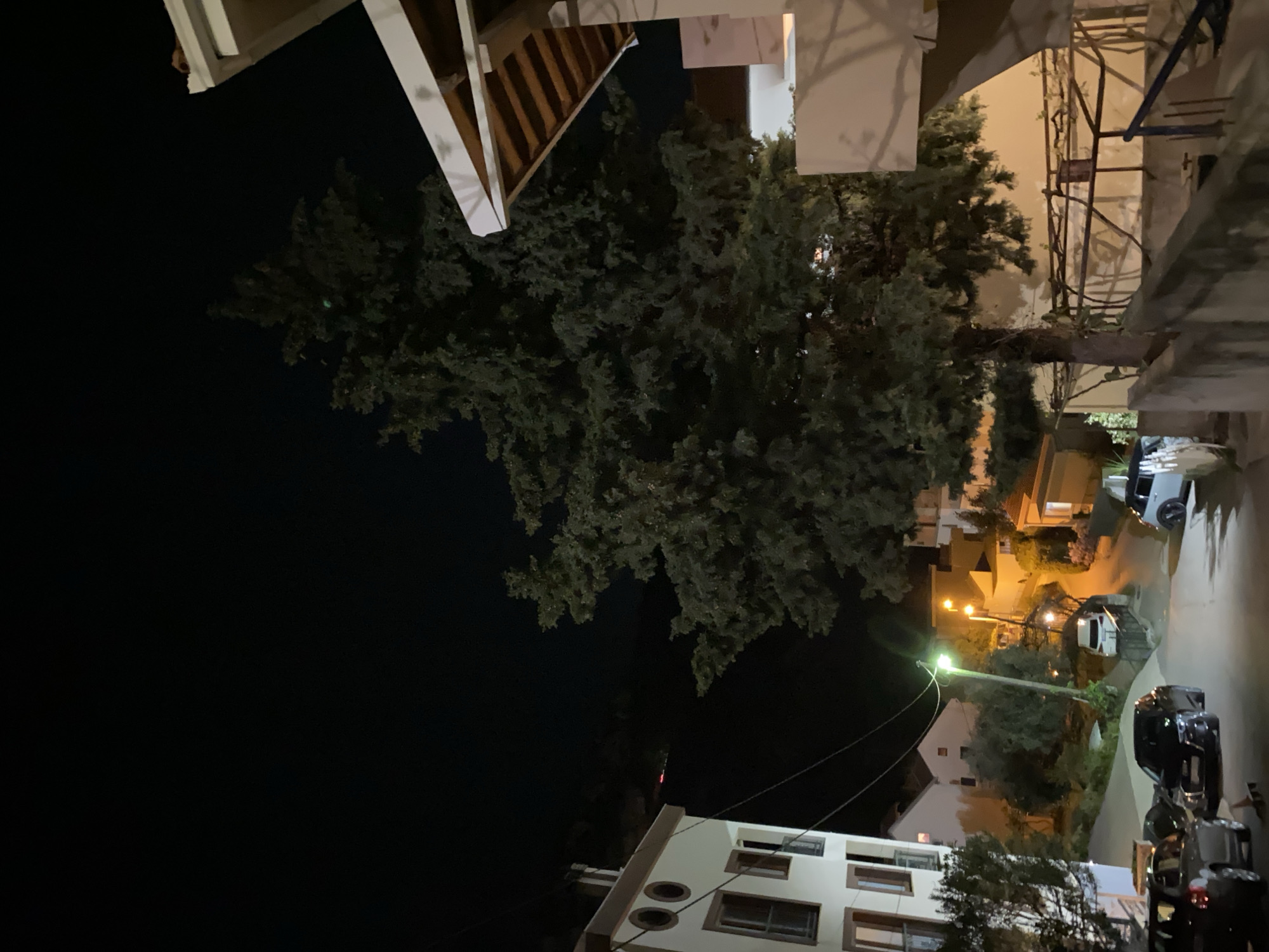 The image depicts a night scene in Budva, Montenegro. In the foreground, there are trees and the corner of buildings, some with visible windows. Cars are parked along a street illuminated by streetlights. The scene is primarily dark, with light emanating from streetlights and building windows. The architecture appears to be a mix of residential and commercial buildings. The focus is on the interplay of light and shadow in the urban environment.