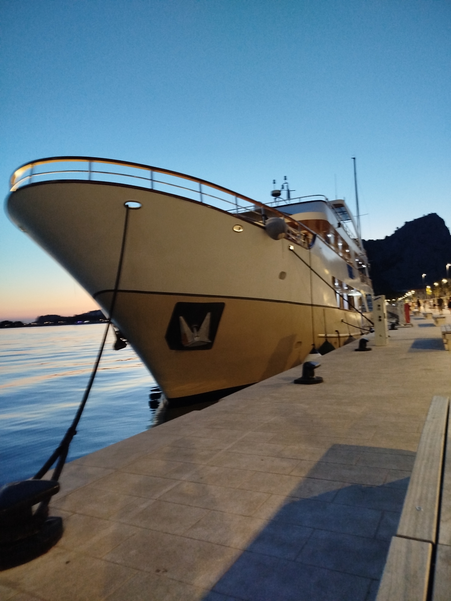 The image captures a large, luxurious yacht docked at a harbor in Omiš, Croatia, during twilight. The sky is a gradient of deep blue to pale orange, suggesting the sun has recently set. The yacht, primarily white and beige, occupies a significant portion of the frame, its bow pointing towards the viewer. It features multiple decks with railings and illuminated windows. The ship is secured to a concrete pier with thick ropes and large bollards. The pier itself is paved with rectangular stones and is illuminated by lampposts along its edge. In the background, a dark, mountainous silhouette looms, characteristic of the Omiš region. The water, reflecting the sky and the yacht's lights, appears calm and rippled. Several people are visible walking along the pier in the distance, adding a sense of activity to the otherwise serene scene. The overall impression is one of tranquility and the end of a day in a coastal Croatian town.