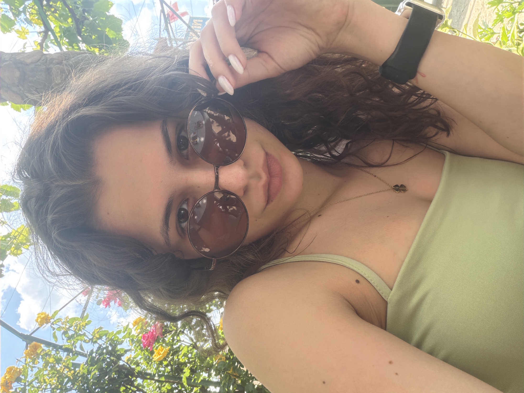 A low-angle, upward-looking selfie captures a young woman outdoors on a bright day. She is seen from the chest up, with her head tilted, looking directly at the camera. Her dark brown, wavy hair frames her face, and she is wearing round, brown-tinted sunglasses that cover most of her eyes. Her lips are slightly parted, suggesting a subtle smile.
She wears a light green sleeveless top or swimsuit. A delicate gold chain necklace with a small, dark clover-shaped pendant hangs around her neck. On her left wrist, she wears a smartwatch with a black band and a metallic casing. Her right hand is raised near her head, with fingers gently touching her hair or the frame of her sunglasses, revealing white-painted nails. Several small moles are visible on her arms and shoulder.
The background is a vibrant mix of nature and sky. Above her, there's a bright blue sky with scattered white clouds, indicative of a sunny day. Around her, lush green foliage with leaves from what appears to be a vine or tree is visible, along with brightly colored flowers in shades of yellow and pink. Reflections in her sunglasses show the bright sky and greenery above. The overall scene conveys a relaxed, summery outdoor setting. There is no visible text.