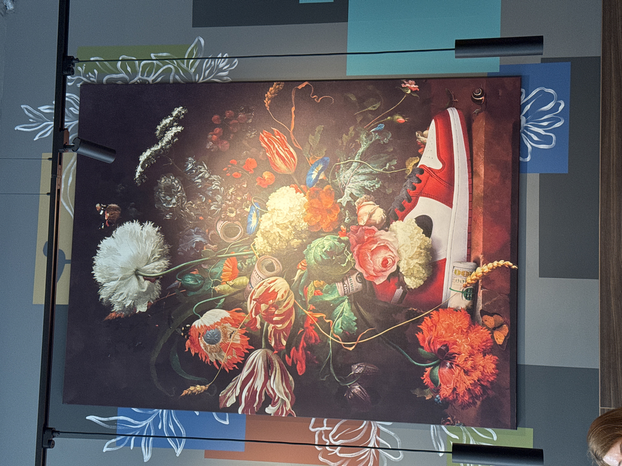 A photograph taken indoors shows a large, framed art piece hanging on a wall. The artwork is a dark, floral still life in the style of the Dutch Masters. However, a modern twist includes a red and white Nike Air Jordan sneaker as a prominent element within the floral arrangement. Currency bills are also placed throughout the piece. The wall behind the artwork has a patchwork design in muted shades of blue, green, gray, and tan, adorned with stylized white floral outlines. Two black track lights, positioned above and below the artwork, illuminate it. Part of a person's head is visible at the bottom right. Given the style of the artwork and interior design, the location is likely an upscale restaurant or gallery. Based on the clues, the location could be in Riga, Latvia.