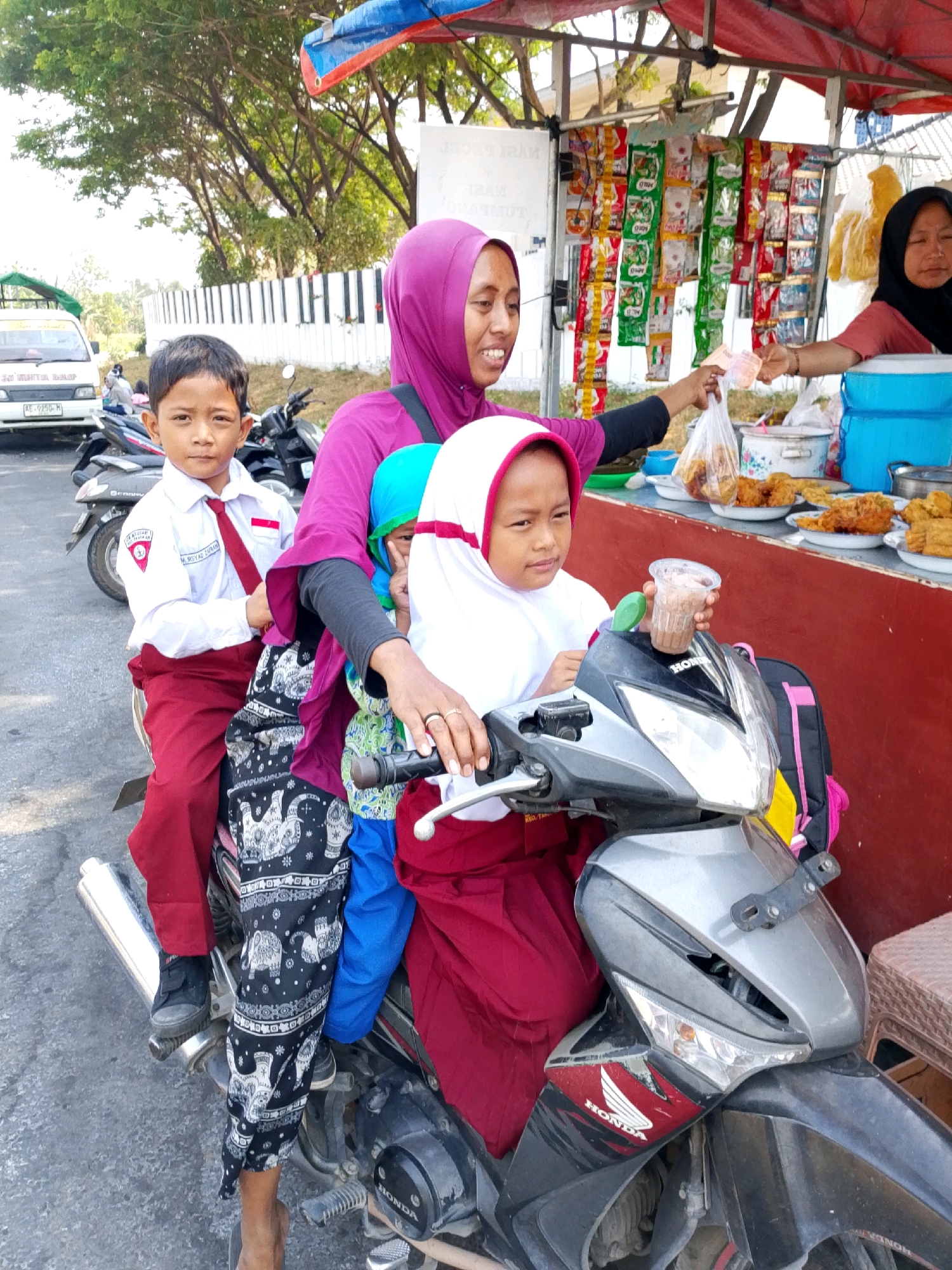 In the city of Bulusari, Indonesia, a mother wearing a purple hijab is riding a motorcycle with three children. The child in front of her, a girl wearing a white hijab, holds a cup with a drink. Behind the mother, a boy in a school uniform with a white shirt and red pants sits between her and another child. The mother is interacting with another woman who is selling goods from a stall. The stall displays various food items and packaged snacks. The scene takes place outdoors, with trees and a white fence visible in the background. A white van is parked nearby.
