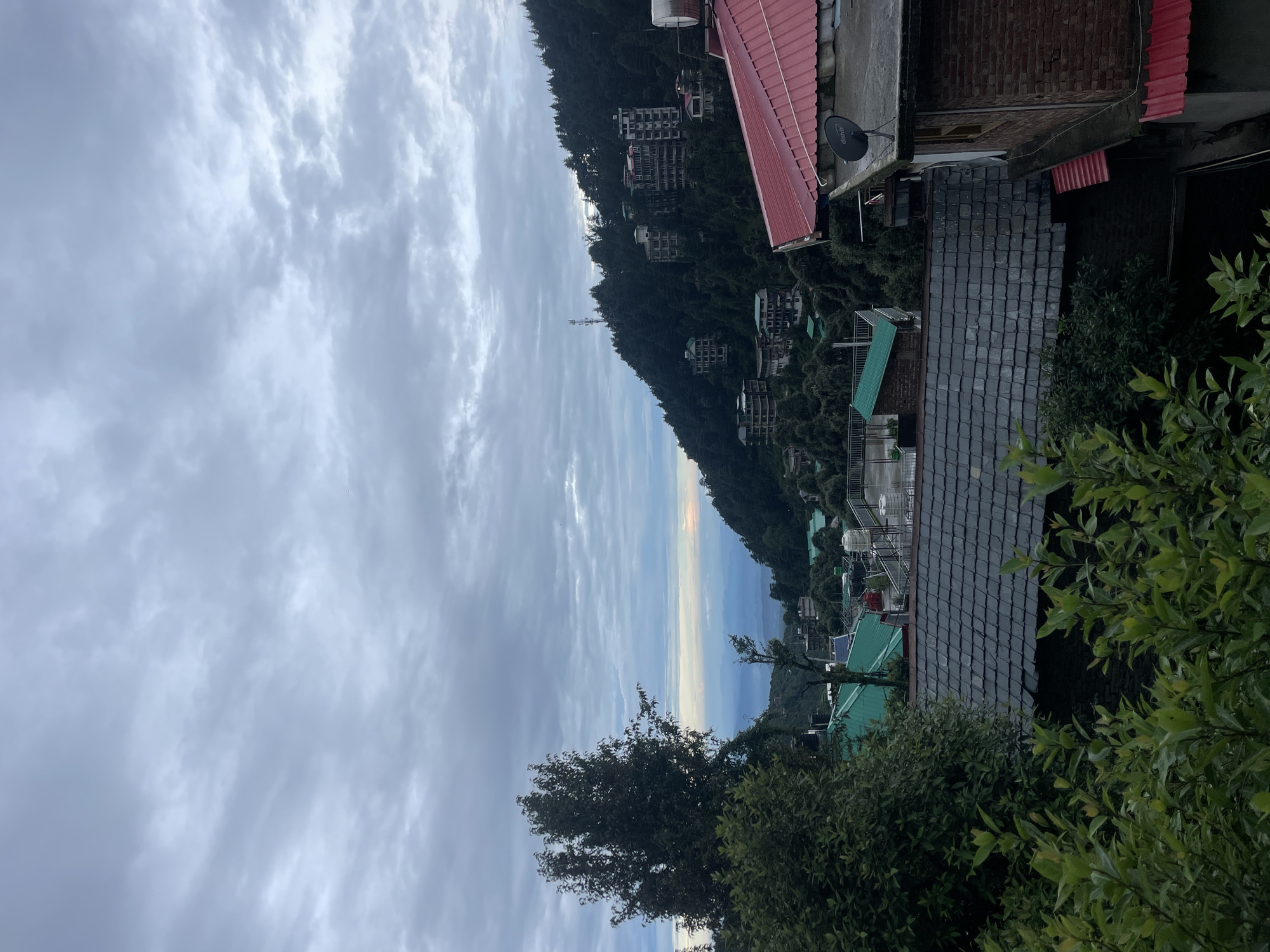 The image depicts a cloudy, overcast sky over a mountainous region, likely Bhagsu, India. In the foreground, lush green foliage frames the bottom of the image. Behind the foliage, a building with a dark, tiled roof is visible. Further back, several buildings with green roofs are scattered among trees. The mid-ground showcases a hillside densely covered with dark green trees, dotted with multi-story buildings. A communication tower stands on the highest point of this hill. In the distance, a hazy view of distant mountains can be seen under the subdued sky. The overall impression is a serene, possibly early morning or late afternoon scene in a hill station.