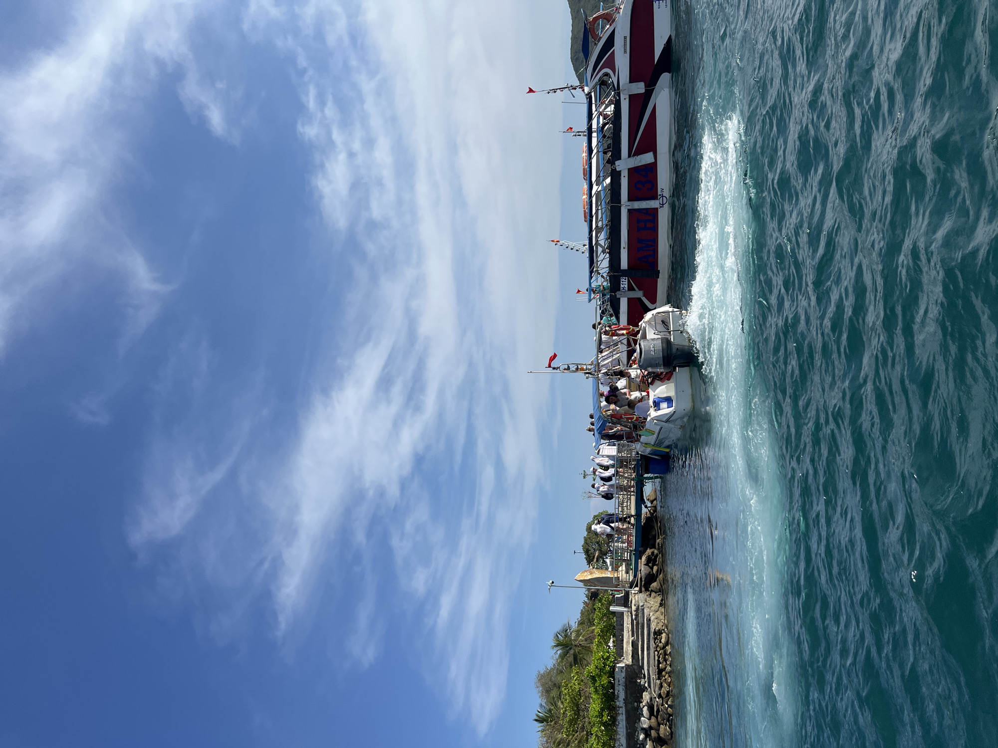 The image shows a view from the city of Nha Trang Ward in Vietnam, looking out at the water where several boats are docked. A large ferry boat with red and white markings is situated beside a smaller white boat. Passengers are visible on both vessels. The water displays churn marks. The sky is blue with sparse clouds. A concrete pier with tropical plants lines the edge of the water in the foreground.