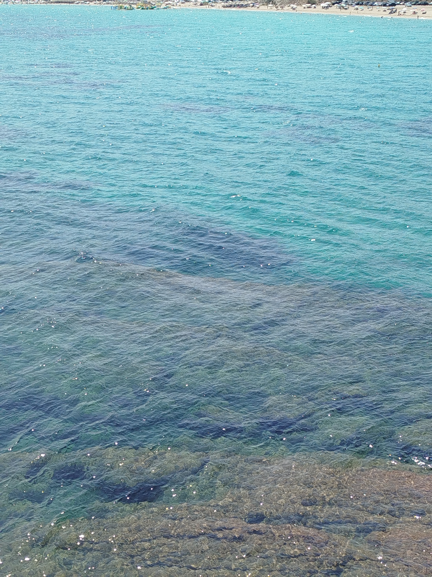 The image captures a view of the Aegean Sea off the coast of Sithonia, Greece. The water transitions from a shallow, clear turquoise near the shoreline to a deeper, darker blue further out. The seabed is visible in the shallower areas, with submerged rocks and sand. Light shimmers on the surface of the water, indicating a sunny day. In the distance, a beach lined with umbrellas and people can be seen, suggesting a recreational area. The overall scene depicts a serene coastal environment.