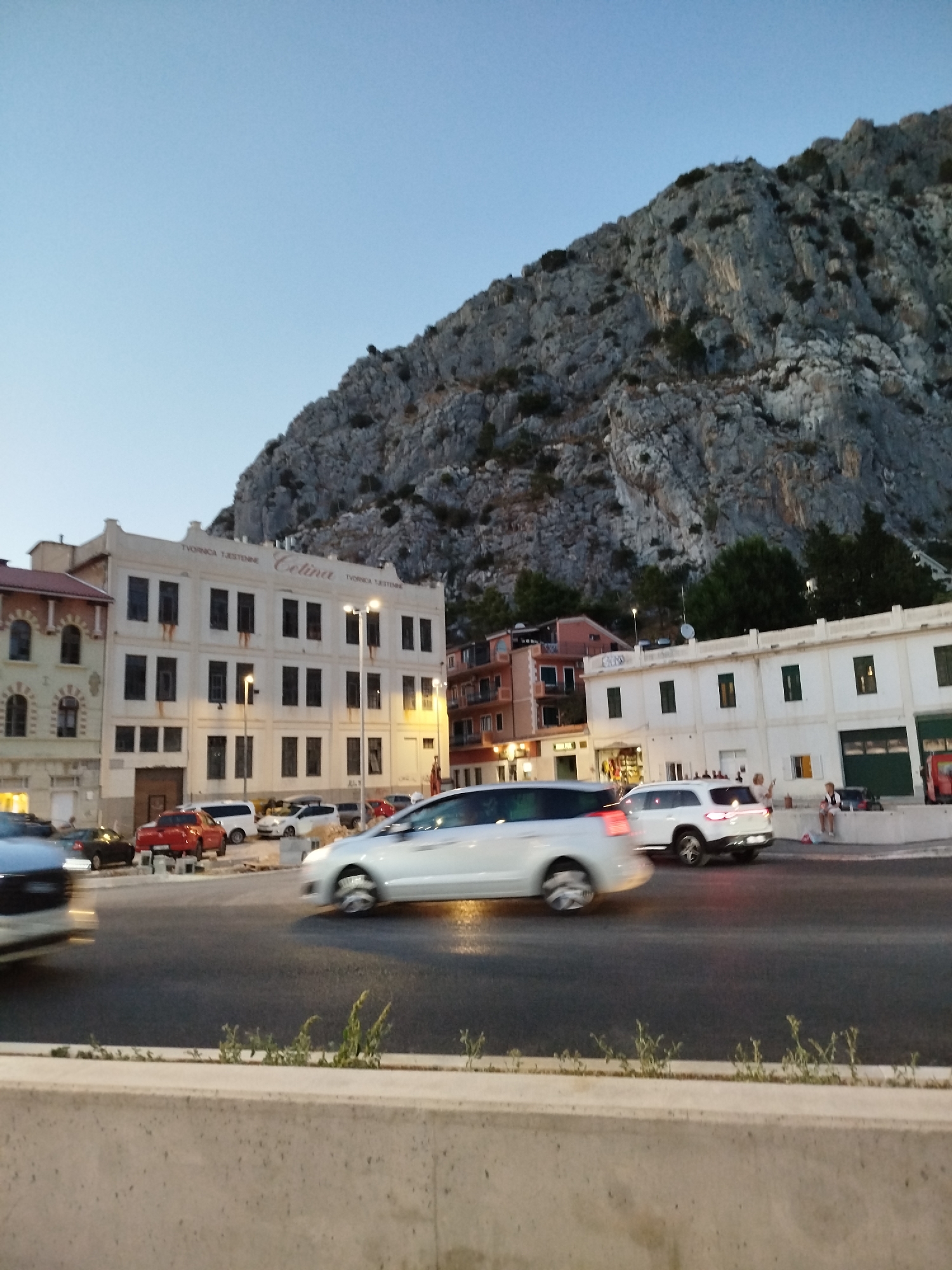 The image shows a view of the city of Omiš, Croatia, with a prominent rocky mountain dominating the background. In the foreground, a road is visible with several cars in motion, some blurred due to movement. A light-colored minivan is in the center, moving left to right. To its left, a red car is parked, and further left, a darker colored car is also parked. A white SUV is visible on the right side of the road, also moving. Beyond the road, buildings are situated. The largest building is a pale cream-colored structure with numerous dark windows and the inscription "TVORNICA TJESTENINE Celina" above the windows. To the left of this building is another building with arched windows. To the right, there are white buildings with green doors or shutters. The sky is a clear light blue, suggesting it is either early morning or late evening. A concrete barrier runs along the bottom of the image.
