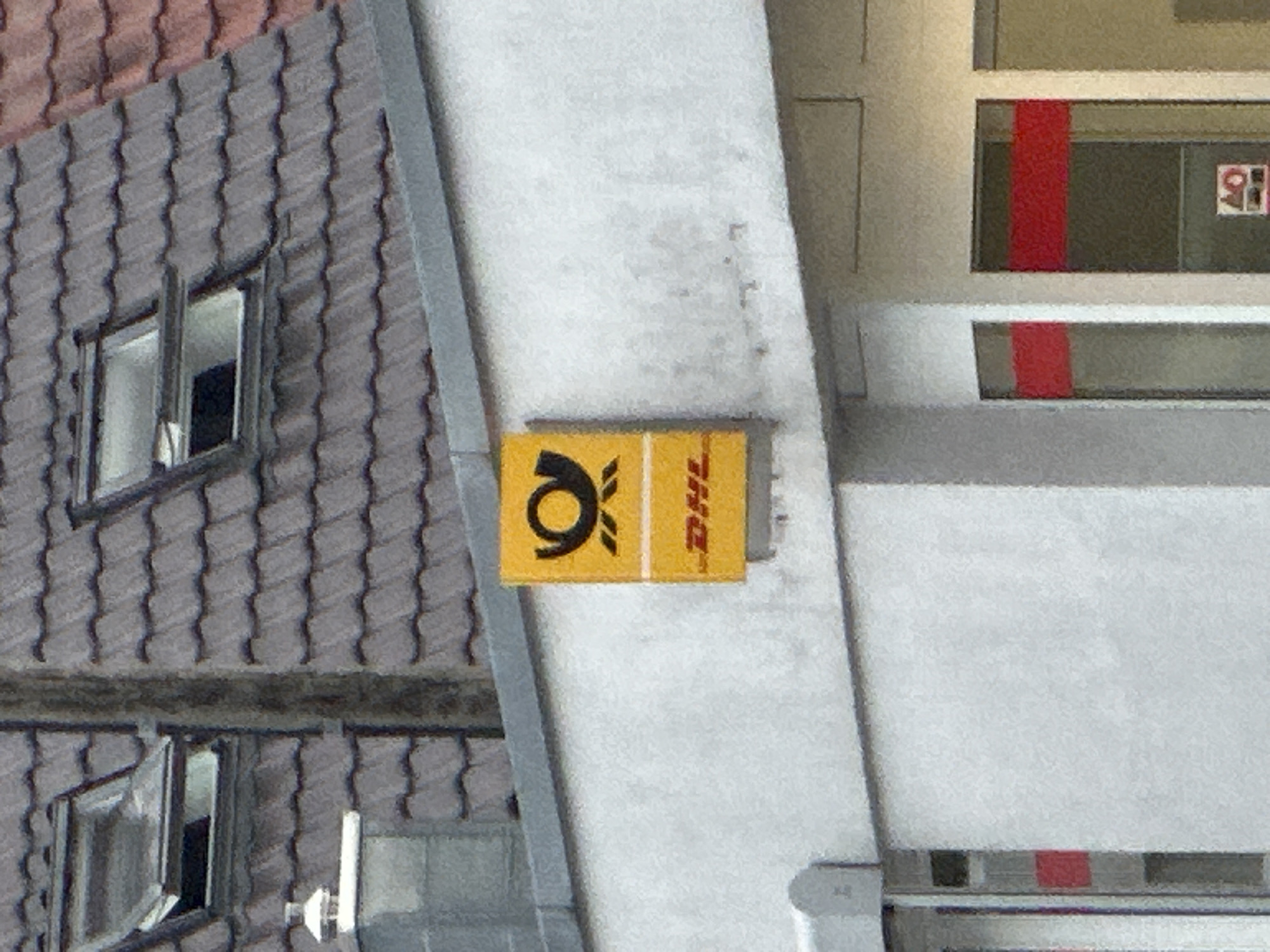 The image shows a yellow sign with black lettering that says "DHL" on a white building. The sign also features a black horn symbol. The building is located in an urban environment, with a grey tiled roof and a dormer window visible in the background. The weather appears to be overcast. There are no people visible in the image.