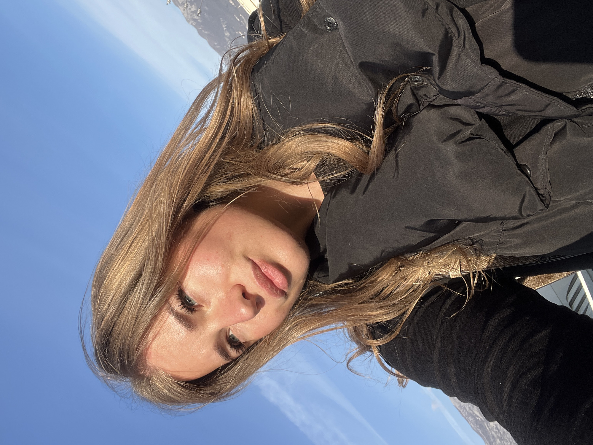A young adult female is depicted in a close-up, high-angle shot, facing towards the upper-left of the frame with her head slightly tilted. She possesses long, wavy, light brown hair extending past her shoulders. Her complexion is fair, with a subtle flush on her cheeks, and her light-colored eyes, featuring prominent eyelashes, are directed upwards and to the left. Her lips are full and slightly parted. She is dressed in a black long-sleeved garment, overlaid with a black puffer vest or jacket, a metallic snap button visible on its front. The background consists of a clear, bright blue sky marked by faint wispy clouds and a distinct white contrail in the upper-left quadrant. Mountains or large hills are visible in the mid-ground on the right and lower-left. A light-colored horizontal structure, possibly a railing or balcony, is partially visible in the bottom-left corner, suggesting an elevated vantage point. The scene is brightly illuminated by direct sunlight. This elevated perspective, featuring mountains and clear skies, is consistent with a scenic viewpoint within or overlooking the coastal city of Budva, Montenegro.