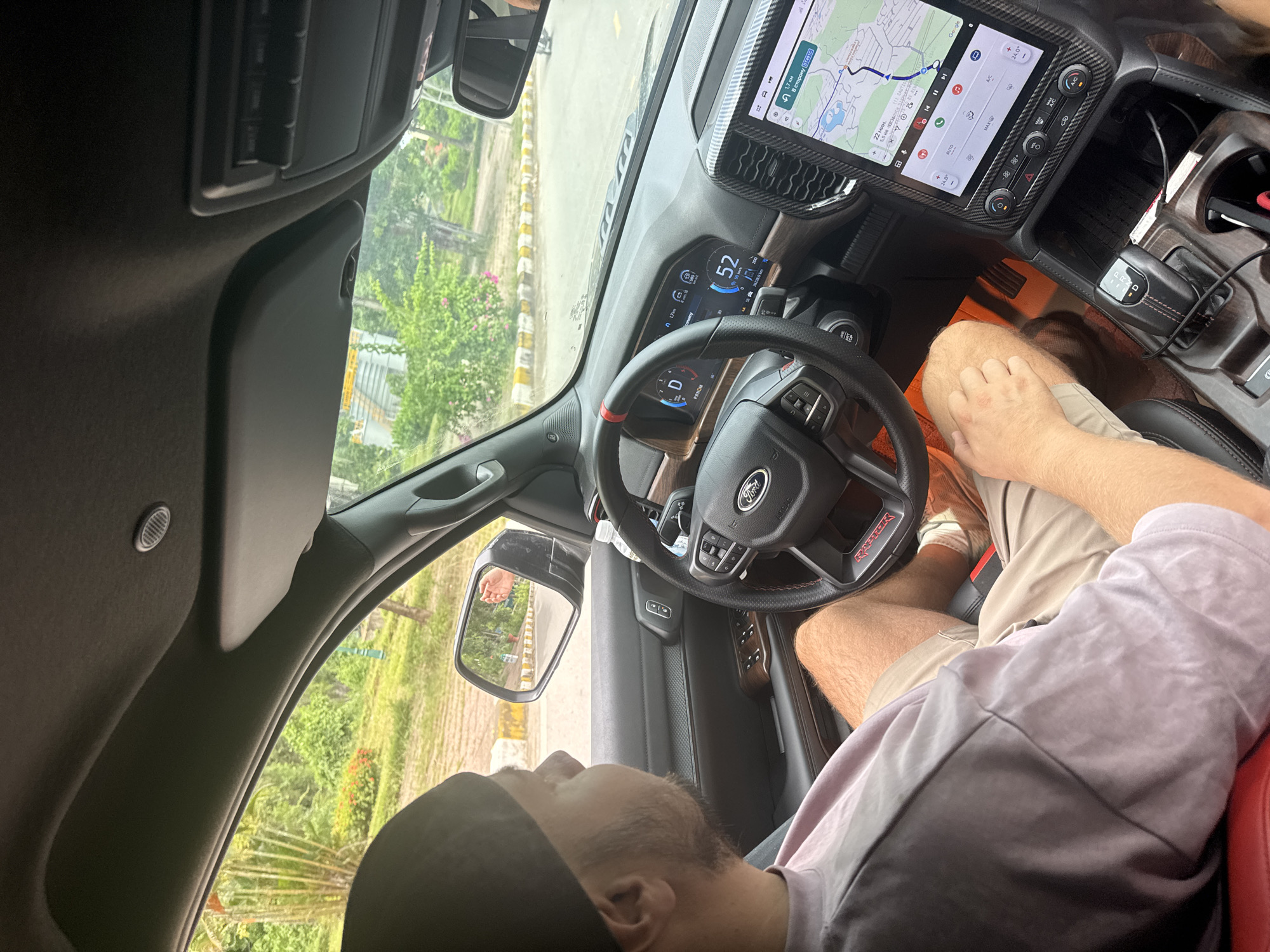 In Nam Hoa Lư Ward, Vietnam, an image captures the interior of a Ford vehicle from a passenger's perspective. A person wearing a dark baseball cap and a light purple t-shirt is seated in the driver's position, looking to the left. The vehicle's dashboard displays a digital instrument cluster showing a speed of 52 km/h and a navigation system on a central touchscreen. The surrounding environment visible through the windshield and side windows appears to be a roadside area with trees and some buildings in the distance. The scene is illuminated by natural daylight, indicating it is daytime.