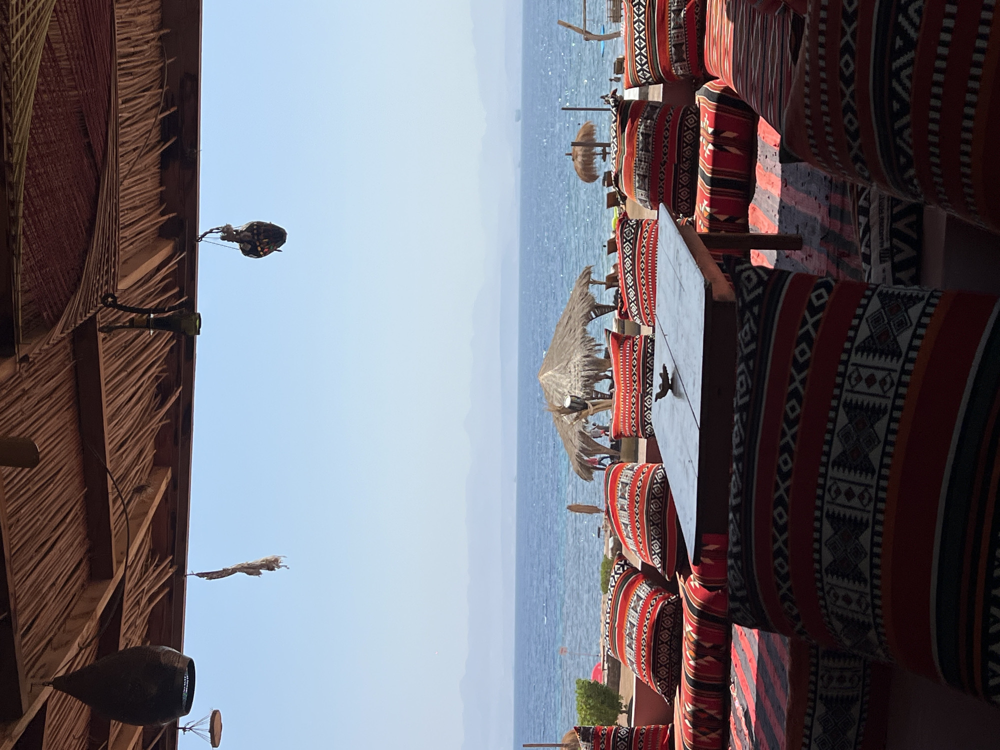 The image depicts an outdoor seating area at a beachfront establishment in Nuweiba, Egypt. The foreground is dominated by low, floor-level seating arrangements covered in cushions with intricate red, black, and white geometric patterns. A rustic wooden table sits in the center of the seating area. Behind the seating, a blue sea stretches to the horizon, where a hazy coastline is visible under a clear, light blue sky. Several thatched-roof umbrellas and lounge chairs are scattered along the sandy beach, suggesting a relaxed atmosphere. The seating area is sheltered by a thatched roof structure, with decorative lamps hanging from it. The overall scene conveys a sense of a casual, beachside dining or lounging experience.