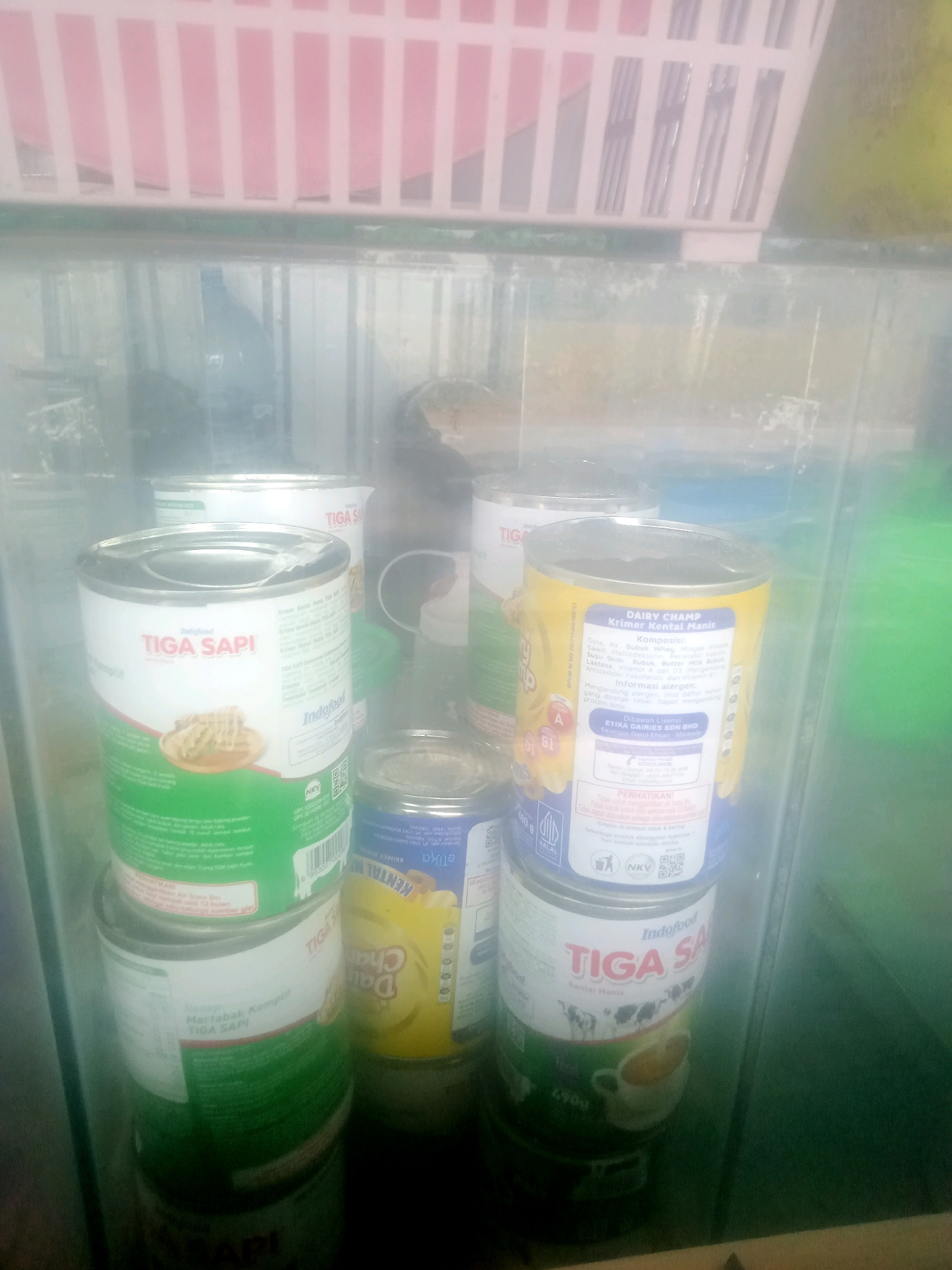 The image shows several cans of condensed milk stacked inside a glass display case, likely in a convenience store or small shop in Penusupan, Indonesia. The cans are mostly Tiga Sapi brand, which is a popular Indonesian dairy product. One can is a yellow Dairy Champ brand. The display case is illuminated, and the background is a pink and white striped rack. The overall lighting is dim, and the glass of the display case is smudged and reflective. There are no people visible in the image. The focus is on the stacked cans of condensed milk.