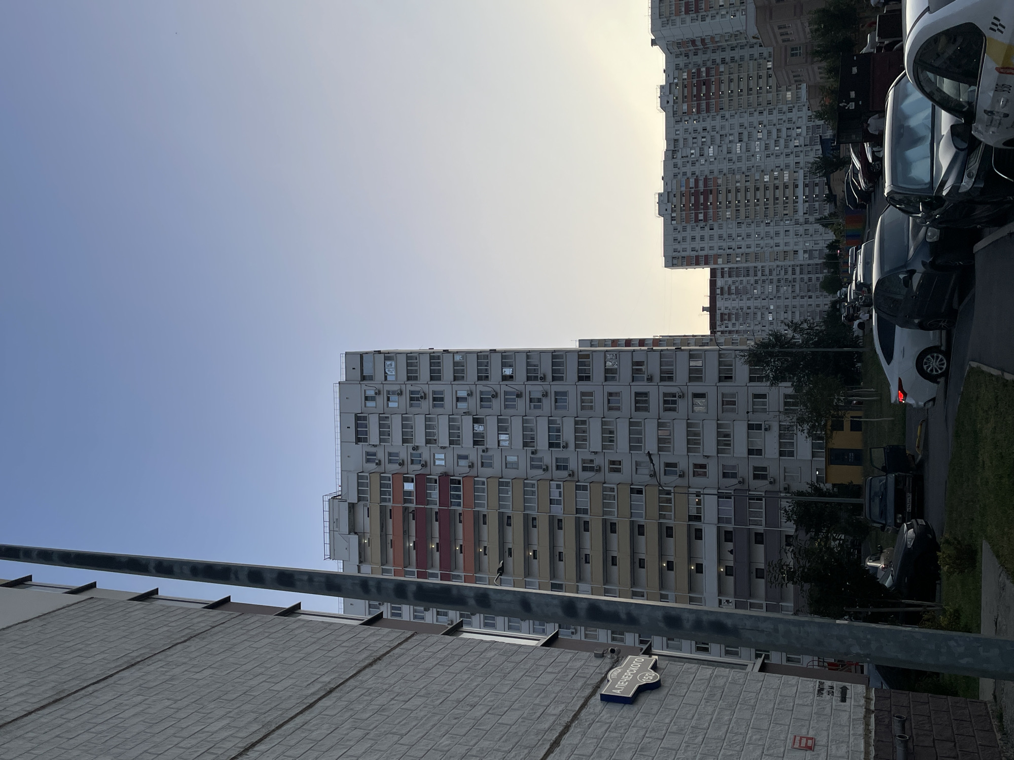 The image shows a street scene in Rostov-on-Don, Russia, likely in a residential area, characterized by multi-story apartment buildings and parked cars.
In the foreground, a grey, textured building wall is visible on the left, with a street sign that reads "А. ПЕЧЕРСКОГО" (A. Pechersky Street) and "15/3". A metal pole, likely a lamppost, runs vertically along the left side of the image. Beyond this, a tall, light-colored apartment building with numerous windows and balconies dominates the mid-ground. This building features bands of orange and red on some of its upper floors. Further into the background, more similar apartment buildings stretch across the horizon, some with visible balconies and windows.
The street level shows several parked cars, including a dark sedan with its driver's side door open. Another dark sedan, a white car, and a taxi with visible yellow and white markings are also present. The environment appears to be a residential courtyard or street, with patches of grass and trees interspersed between the buildings and cars. The sky is clear and light blue, transitioning to a paler hue near the horizon, suggesting it is either early morning or late afternoon. There are no visible people actively engaged in interactions, though a figure can be seen on a balcony of the central apartment building.