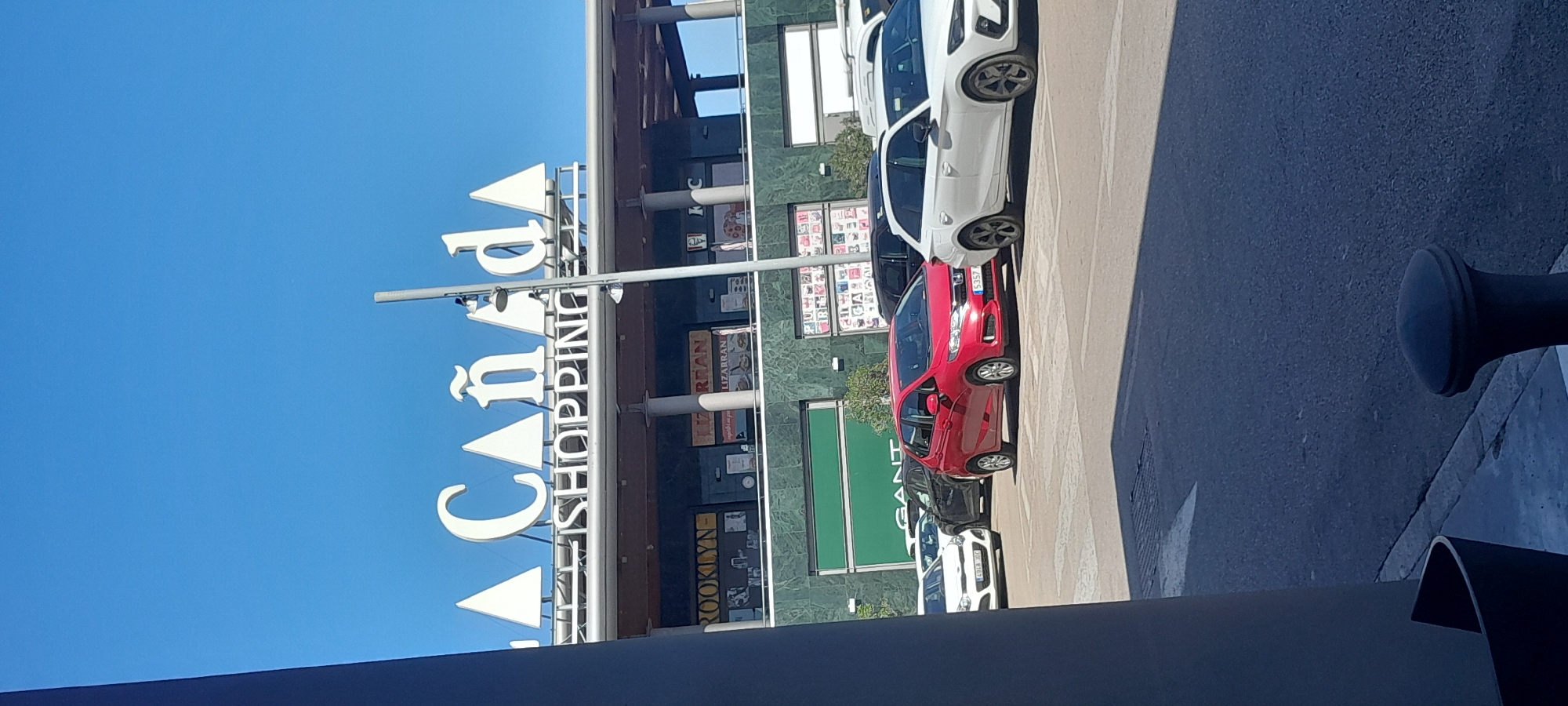 The image captures a sunny daytime scene outside a modern shopping center. The main subject is a large, multi-level commercial building on the left, featuring a facade with dark green or black panels and reflective surfaces. A prominent vertical sign affixed to the building clearly displays "CAÑADA SHOPPING" in large, white, capital letters.
Several store signs are visible on the building's various levels, including "KFC," "LIZARRAN," "BROOKLYN," and a partially visible "GANT" logo. Above and to the left of the building, the sky is a clear, bright blue, indicating good weather.
On the right side of the image, a paved parking area or road runs parallel to the shopping center. Several cars are parked here, facing the building. A white hatchback is visible in the foreground, followed by a bright red hatchback with a partially visible license plate showing "5357". Another white car is parked further down, with a license plate containing "493...HGK." Strong shadows are cast across the pavement, suggesting direct sunlight. In the bottom right corner, a dark, cylindrical bollard or pole is visible. No people are present in the frame, and the scene conveys a calm atmosphere typical of a commercial area.