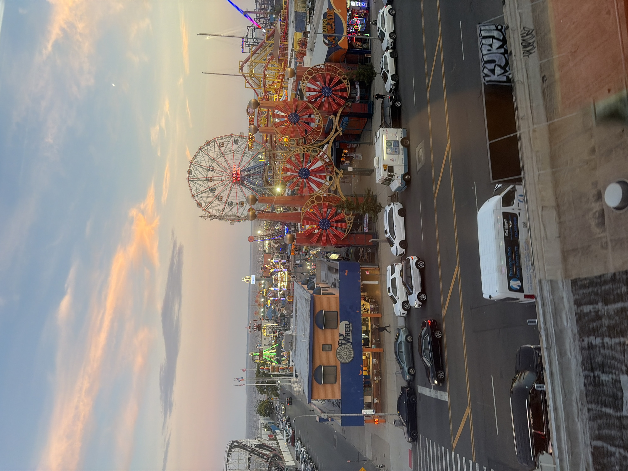 This is an eye-level, medium shot of Coney Island Amusement Park in Brooklyn, New York City, USA, during a twilight hour, with the sky displaying soft pastel colors. The scene is dominated by amusement park attractions, including a large Ferris wheel labeled "Wonder Wheel" and a roller coaster. In the foreground, a street runs horizontally across the image, with several cars parked and driving on it. On the left side of the street, there is a building with orange walls and a blue awning that reads "FLY WHEEL CAFE". Further into the park, there are more rides and structures, and the ocean is visible in the background. The lighting suggests it is either sunset or sunrise, with the artificial lights of the park beginning to illuminate the attractions.