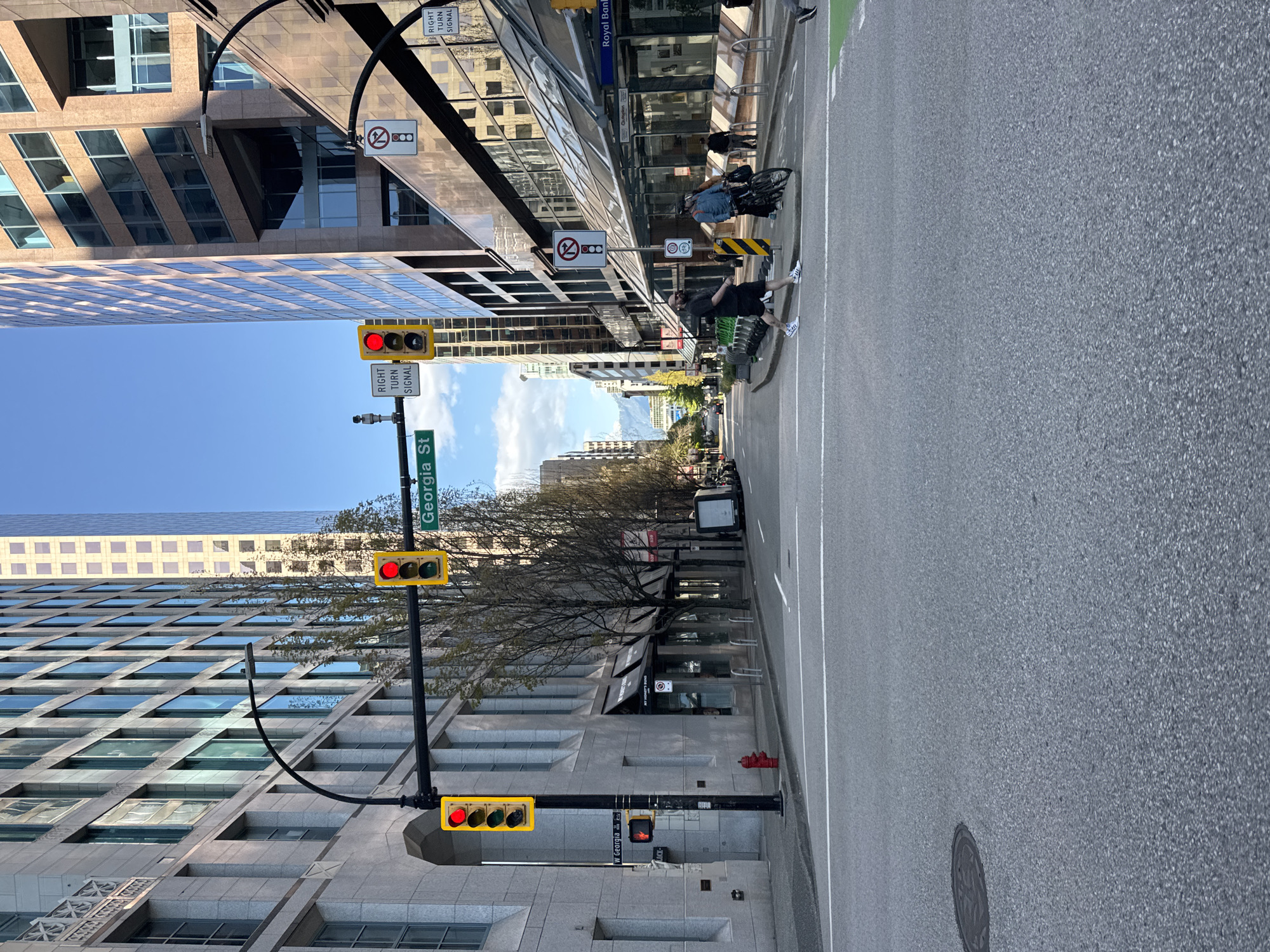 The image depicts a street scene in Vancouver, Canada, specifically along Georgia Street, as indicated by a street sign. The perspective is looking down the street, with buildings lining either side. Pedestrians and a cyclist are visible on the right side of the road, with one pedestrian appearing to be in the process of crossing. Traffic lights are positioned along the street, displaying red signals at multiple points. The sky is blue, with scattered clouds. The street itself is paved, with lane markings and a manhole cover visible. The environment suggests an urban setting with commercial buildings.