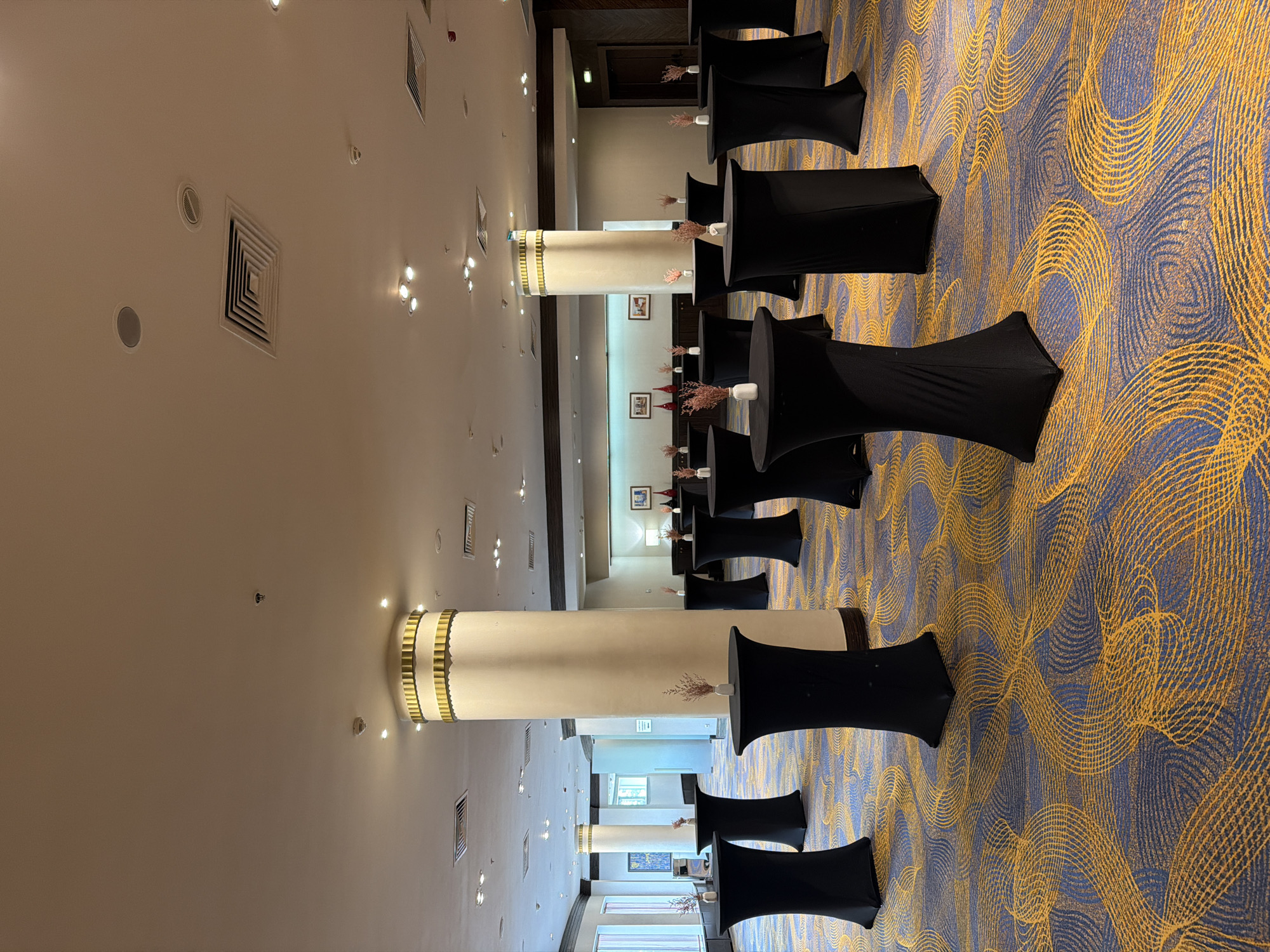 The image shows an interior space, possibly a large ballroom or conference hall, featuring several high cocktail tables draped in black fabric. The tables are arranged in rows along the right side of the room. A large column stands in the middle of the hall, and the flooring is patterned with a blue and gold swirl design. The walls are light-colored, and ceiling lighting is visible. No people are clearly visible in the image. The location is unknown, so I cannot provide the requested city information.