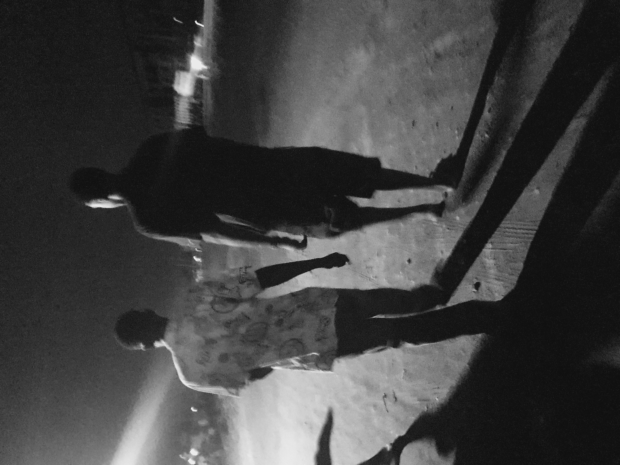 A black and white photograph depicts two people walking on a paved surface, possibly a street or sidewalk, in Lokoja, Nigeria. The image is oriented with the subjects walking from right to left. The person closest to the camera is wearing a patterned shirt and long pants. The other person is wearing a dark T-shirt and shorts. The lighting is dim and creates long, distinct shadows behind the subjects. In the background, there are blurred structures and lights, suggesting a cityscape. The photo has a grainy texture.