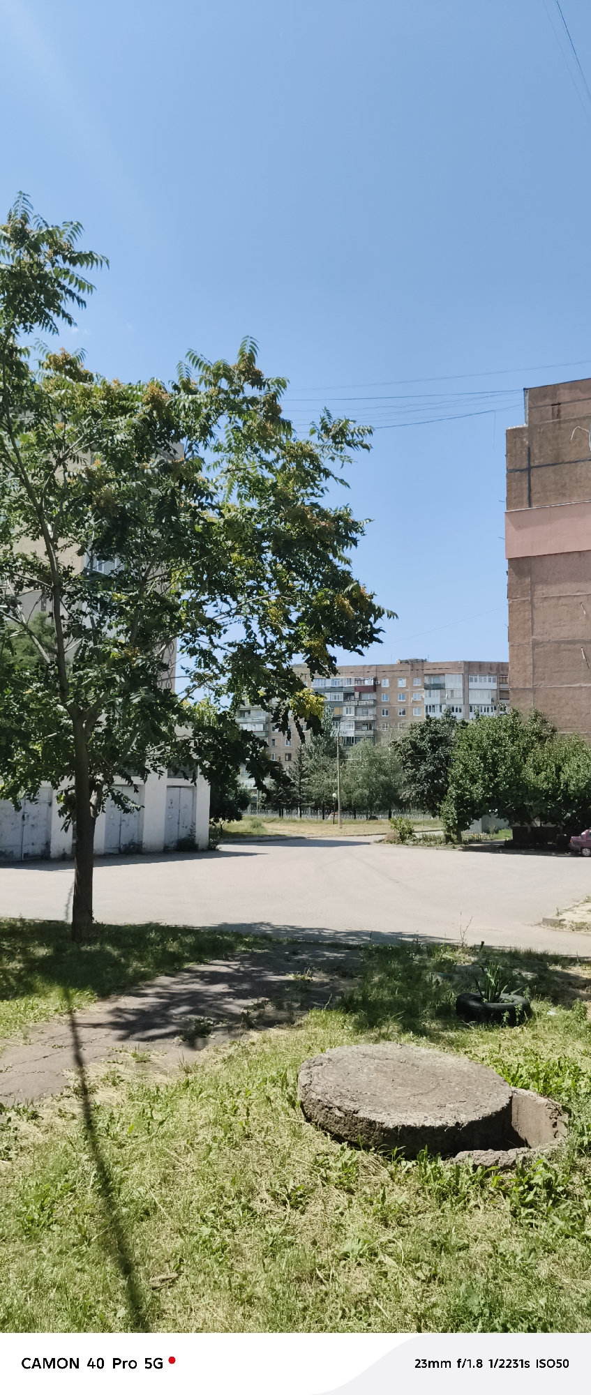 The image depicts an outdoor scene in the city of Kramatorsk, Ukraine, under a clear blue sky. In the foreground, a patch of grass is visible, with a circular concrete slab, likely a manhole cover, partially embedded in it. A thin tree trunk casts a shadow across the grass and a paved area. Beyond this, a paved lot or courtyard is visible, bordered by a low white building with several doors on the left. Further in the background, a line of trees separates the courtyard from a row of apartment buildings. The buildings are multi-story structures with visible windows and balconies, characteristic of Soviet-era architecture. A large tree with green leaves and clusters of small yellowish-brown fruits or seeds is prominently featured on the left side of the frame, obscuring parts of the buildings. The overall environment suggests a residential area within the city. The bottom edge of the image displays text that reads "CAMON 40 Pro 5G" and "23mm f/1.8 1/2231s ISO50", indicating it is a photograph taken with a specific camera model and settings. There are no people visible in the image, and no discernible actions or interactions are taking place. The scene is static and presents a view of urban infrastructure and vegetation.