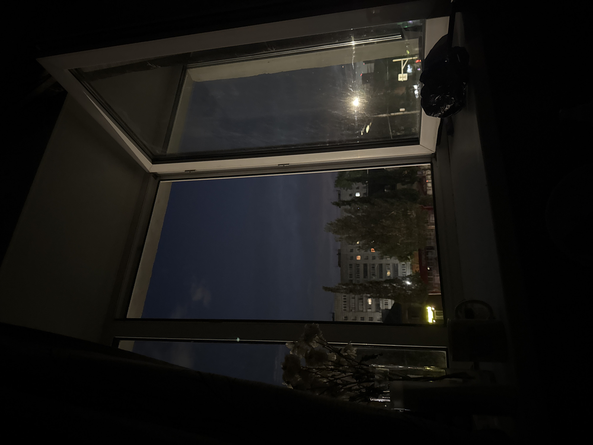 An open window provides a view of a night scene in Perm, Russia. The view includes a multi-story building with illuminated windows, suggesting it is either an apartment complex or a commercial building with late-night operations. Trees partially obscure the building. Above, the sky is visible through the window, indicating dusk or night. The presence of a street light in the top section of the window suggests the view is towards a street or road. The interior of the room is dark, but some objects can be discerned near the window, including what appears to be a bouquet of flowers and an indistinct object to the right. A black plastic bag is hanging on the right side of the window frame.