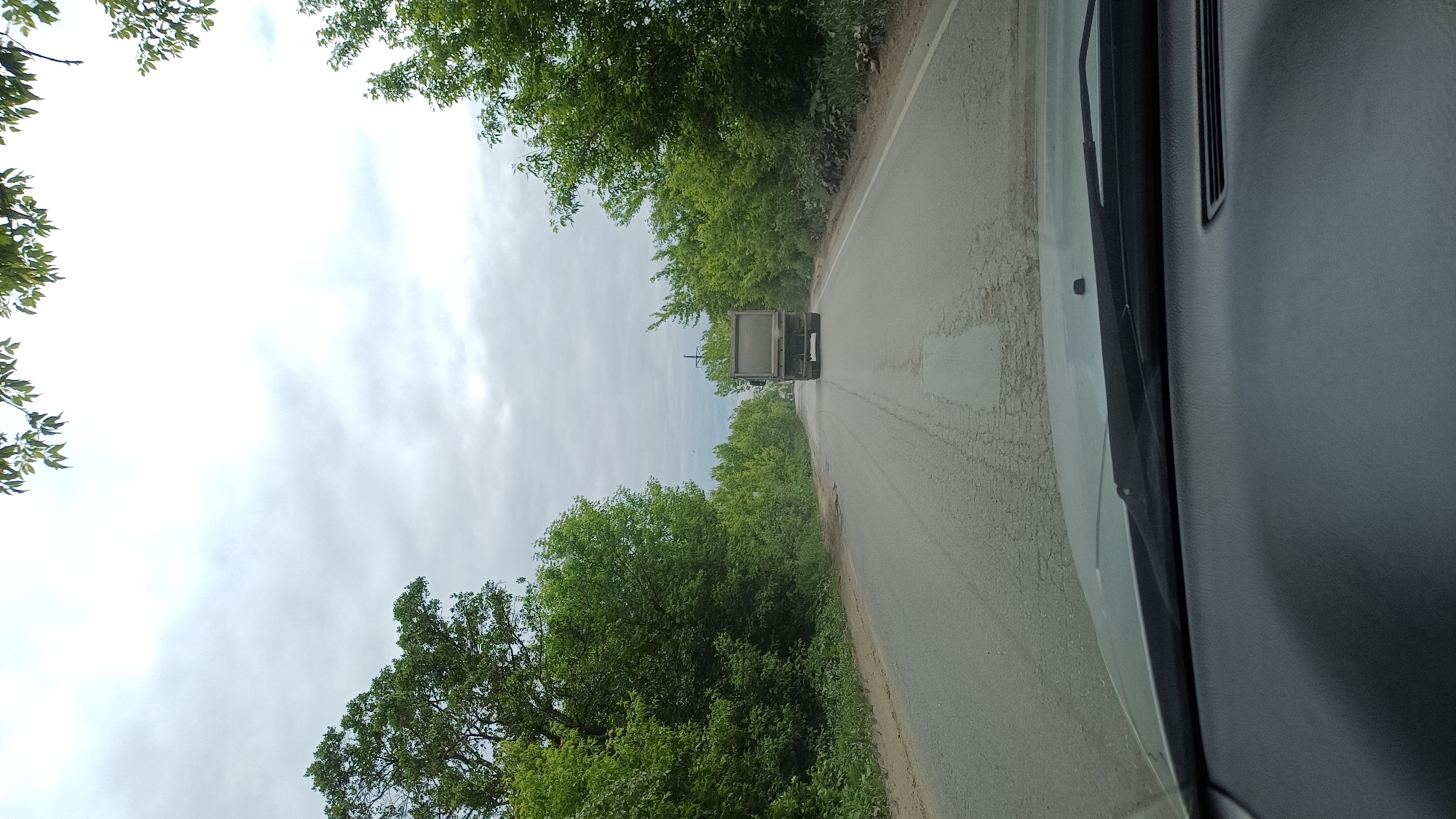The image, captured from the interior of a vehicle, displays a forward-facing view of a paved road. The road surface shows signs of wear, including cracks and tire marks, and is flanked on both sides by dense green trees and foliage, suggesting a rural or semi-rural environment. Ahead, a dark-colored truck is seen driving in the same direction. Behind the truck, a tall utility pole or similar structure is visible against the sky. The sky is largely overcast, indicating cloudy daytime weather. On the right side of the frame, a portion of the vehicle's interior is visible, including what appears to be a dashboard or door panel with an air vent. There are no people visible. The context for this location is Staraya Kupavna, Russia.