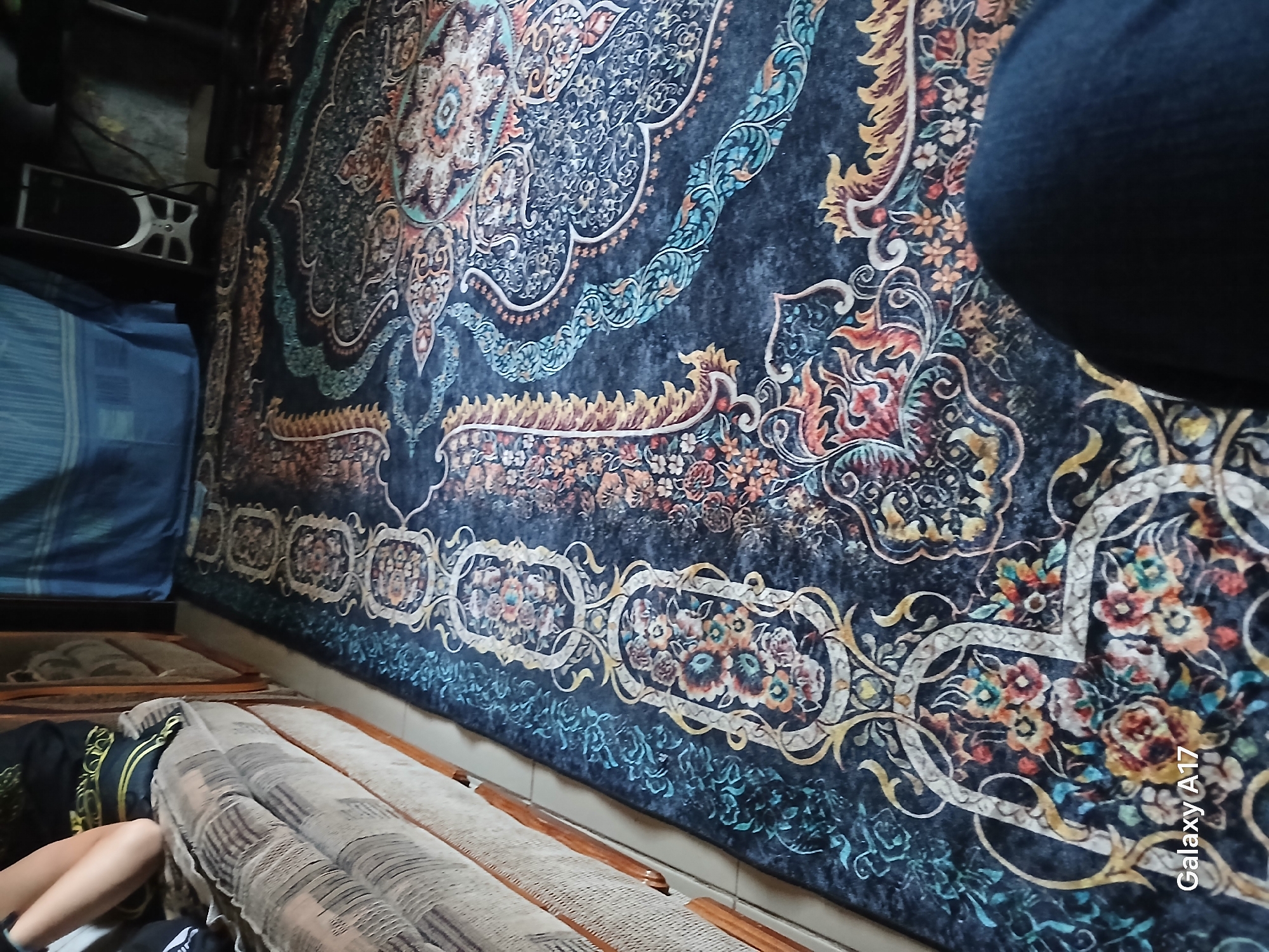 The image shows an interior scene with a person's legs and a portion of their torso visible on the left, seated on a couch. The person is wearing a black shirt with gold embellishments and what appear to be checkered pants or leggings. The couch is upholstered in a patterned fabric in neutral tones. A dark-colored area rug with an ornate, colorful floral and paisley pattern covers a significant portion of the floor. In the background, a blue curtain is visible, along with a computer tower and other objects suggesting a domestic or office environment. The image quality is average, with some blurriness and a watermark that reads "Galaxy A17" in the lower left corner. The overall scene depicts a person relaxing on a couch in a room with a decorative rug. The location within the city of Псыкод, Russia, is not discernable from the visual information provided in the image.