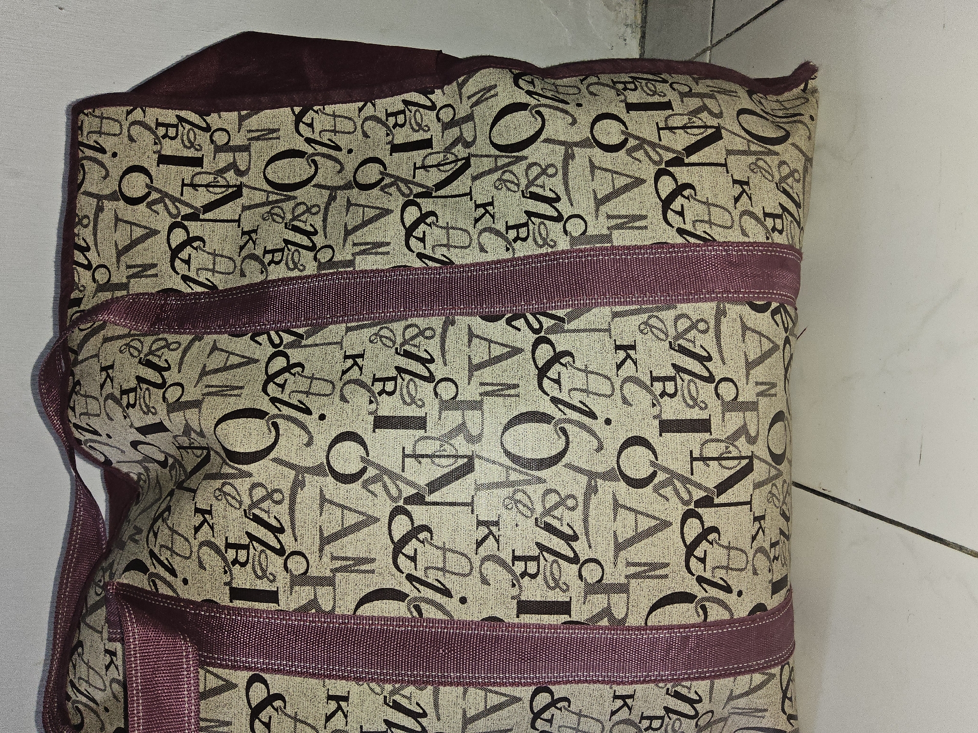 The image shows a close-up, eye-level, vertical shot of a large, tan duffel bag with a repeating pattern of black and dark red letters. The bag has two dark red fabric straps running vertically down the front and two darker red fabric handles at the top. The background is a white wall and a tiled floor, suggesting an indoor setting. The lighting is even, with no strong shadows. The scene does not depict any people, actions, or interactions. The location in Payagaboh, Indonesia is not identifiable from the visual elements present in the image.
