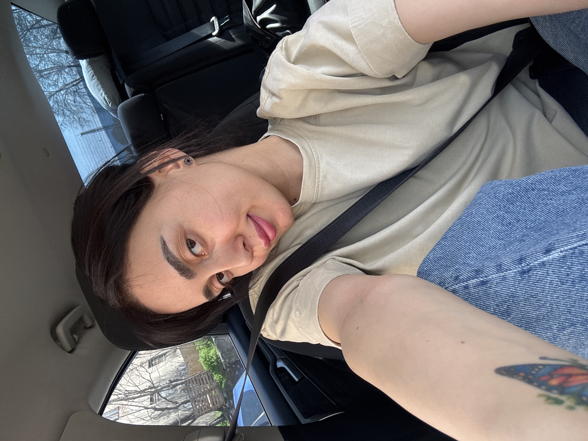 The image shows a young woman in a car. She is wearing a tan t-shirt, blue jeans, and a seatbelt. She has dark hair and is looking at the camera. There is a butterfly tattoo visible on her left arm. Outside the car window, a stone building with bare trees is visible. Based on the architectural style of the building, the location appears to be Tbilisi, Georgia.