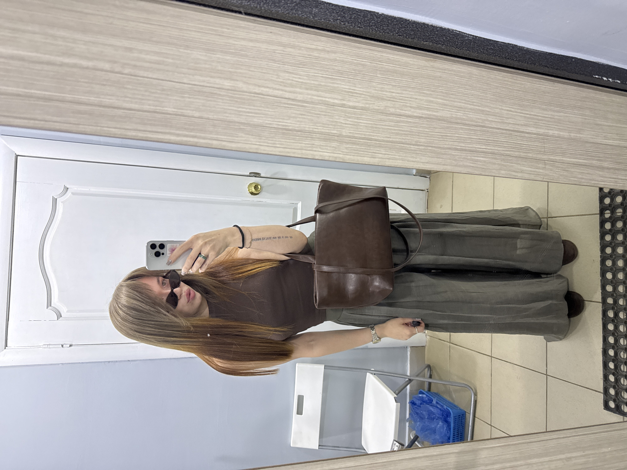 A woman is taking a selfie in a mirror. She is wearing sunglasses, a brown top, and green pants. She has a brown purse over her shoulder and a ring on her finger. She is holding a phone in her left hand. The background includes a white door, a gray wall, a chair, and a blue basket. The location is unknown, with no indication of Chaikovsky, Russia.