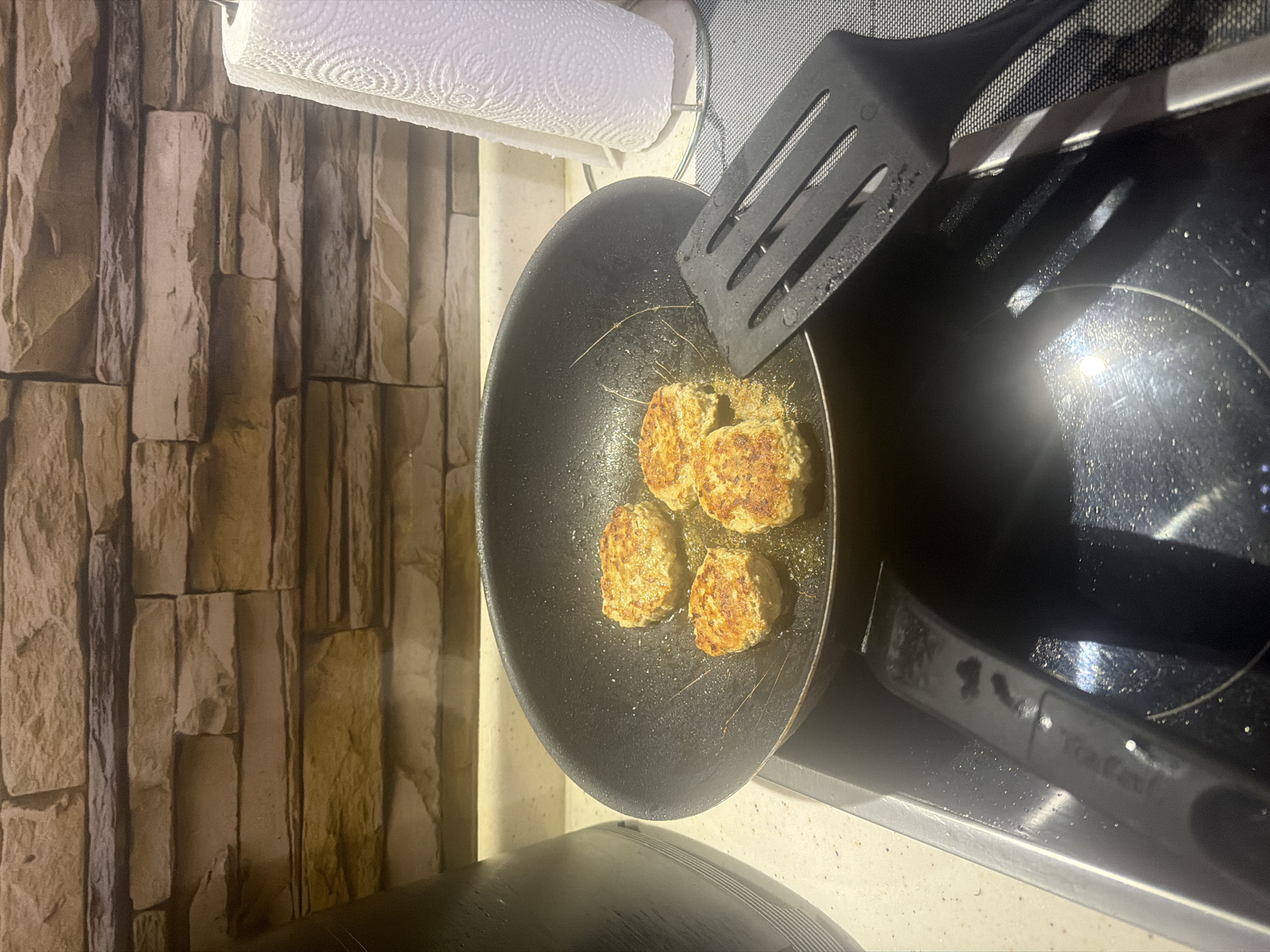The image captures a close-up, top-down view of a kitchen scene featuring food being cooked. A black frying pan, positioned on what appears to be a black induction stovetop, contains four golden-brown, irregularly shaped patties that are frying in oil. A black slotted spatula rests on the edge of the pan, extending over the stovetop.
To the left, a wall covered with decorative tiles or wallpaper resembling stacked, rectangular stone blocks in varying shades of brown and beige forms the background. Above the pan, a white roll of paper towels is held by a metallic dispenser. The stovetop is glossy black, reflecting light and showing scattered splashes or crumbs. Faint, difficult-to-read text, possibly a brand name, is visible on the handle of the frying pan. Notably, several thin, dark strands, resembling hairs or fibers, are present in the pan amidst the cooking patties. The lighting suggests an indoor setting, likely illuminated by artificial light.