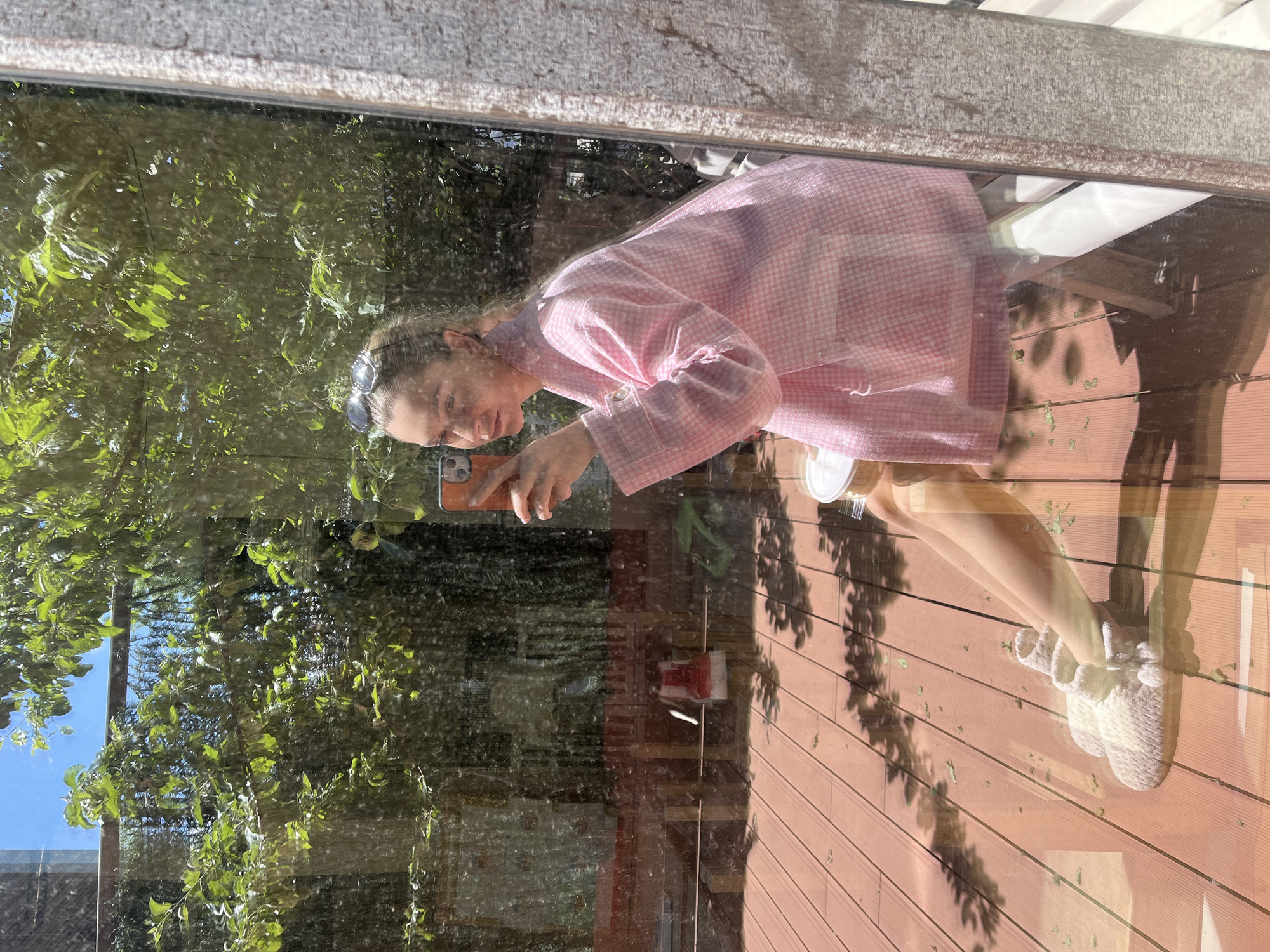 A person is taking a picture in a window. The subject appears to be female, wearing a pink and white gingham robe and white slippers. A phone is being held up by the person to take the photo. Reflections in the window show trees, sky, and wood siding of a building. There is no indication of the specific city.