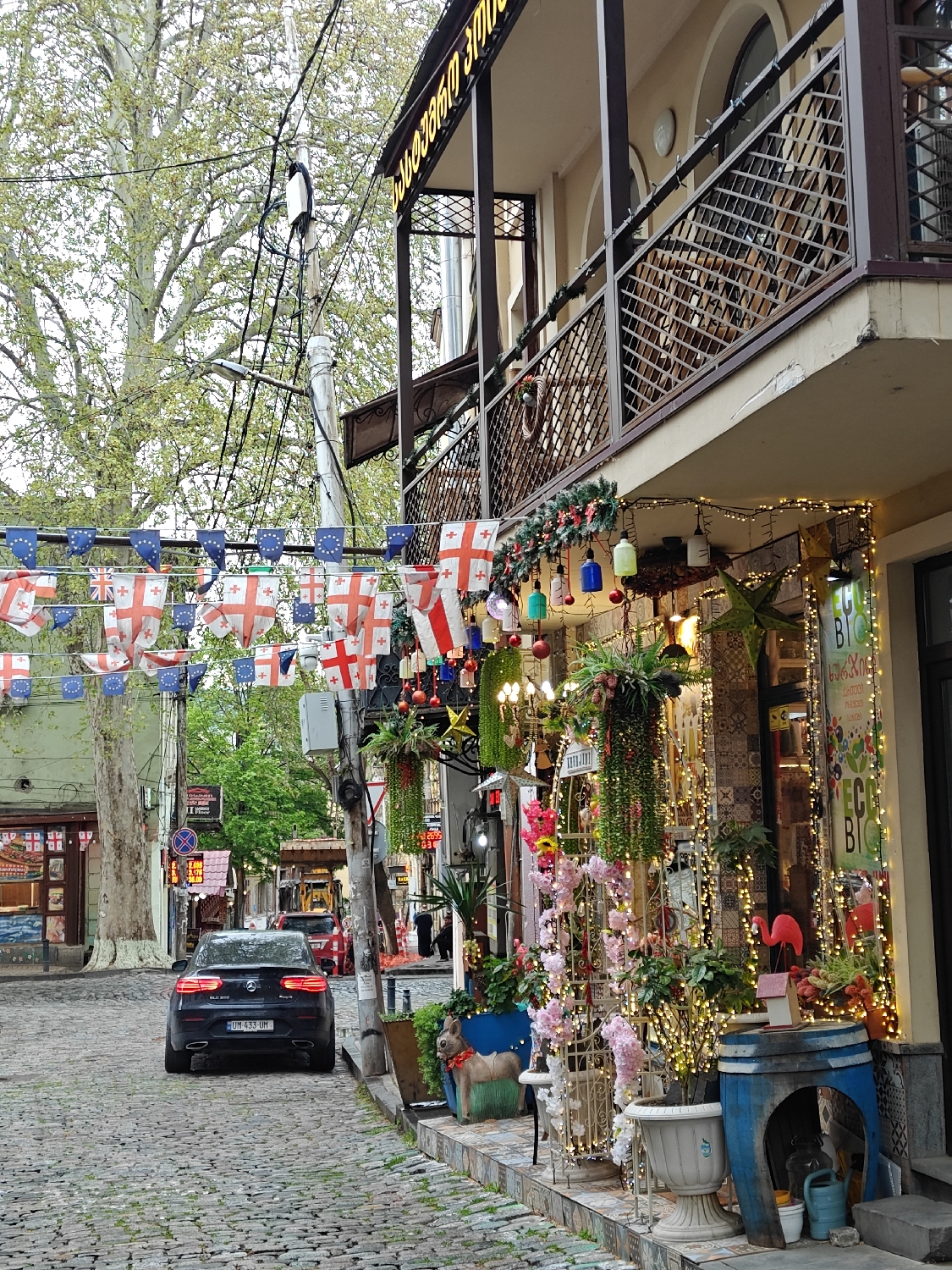 The image presents a vibrant street scene in Tbilisi, Georgia, characterized by its cobblestone path and decorative elements. In the foreground on the right, a storefront is elaborately adorned with numerous string lights, illuminating hanging plants, colorful bells, red ornaments, and artificial pink flowers on trellises. Various potted plants, a small blue barrel, a watering can, a pink flamingo ornament, and a small ceramic dog-like statue further enhance the festive display. A vertical sign on the storefront partially reveals "BIO."
Above this decorated shop, a traditional building features a prominent balcony with detailed metalwork. A sign in Georgian script, "პასაჟი ზამთარი" (Pasazhi Zamtari), translating to "Winter Passage" or "Winter Arcade," is visible along the top of the building.
Strung across the street, colorful bunting displays flags of Georgia (white with a large red cross and four smaller crosses), the European Union (blue with yellow stars), and the United Kingdom (Union Jack), contributing to a lively, international atmosphere.
On the cobblestone street, a black Mercedes-Benz GLC 300 is parked, facing away from the camera. Its Georgian license plate, "UM 433 UM," is clearly visible. In the midground and background, more traditional buildings with balconies are visible, interspersed with large trees featuring light green foliage. Power lines and poles crisscross the upper part of the scene.
The image captures a daytime setting, likely with mild weather, emphasizing the decorative and welcoming nature of this area in Tbilisi. No specific human activity is depicted, but the scene suggests a bustling, possibly tourist-oriented, street.