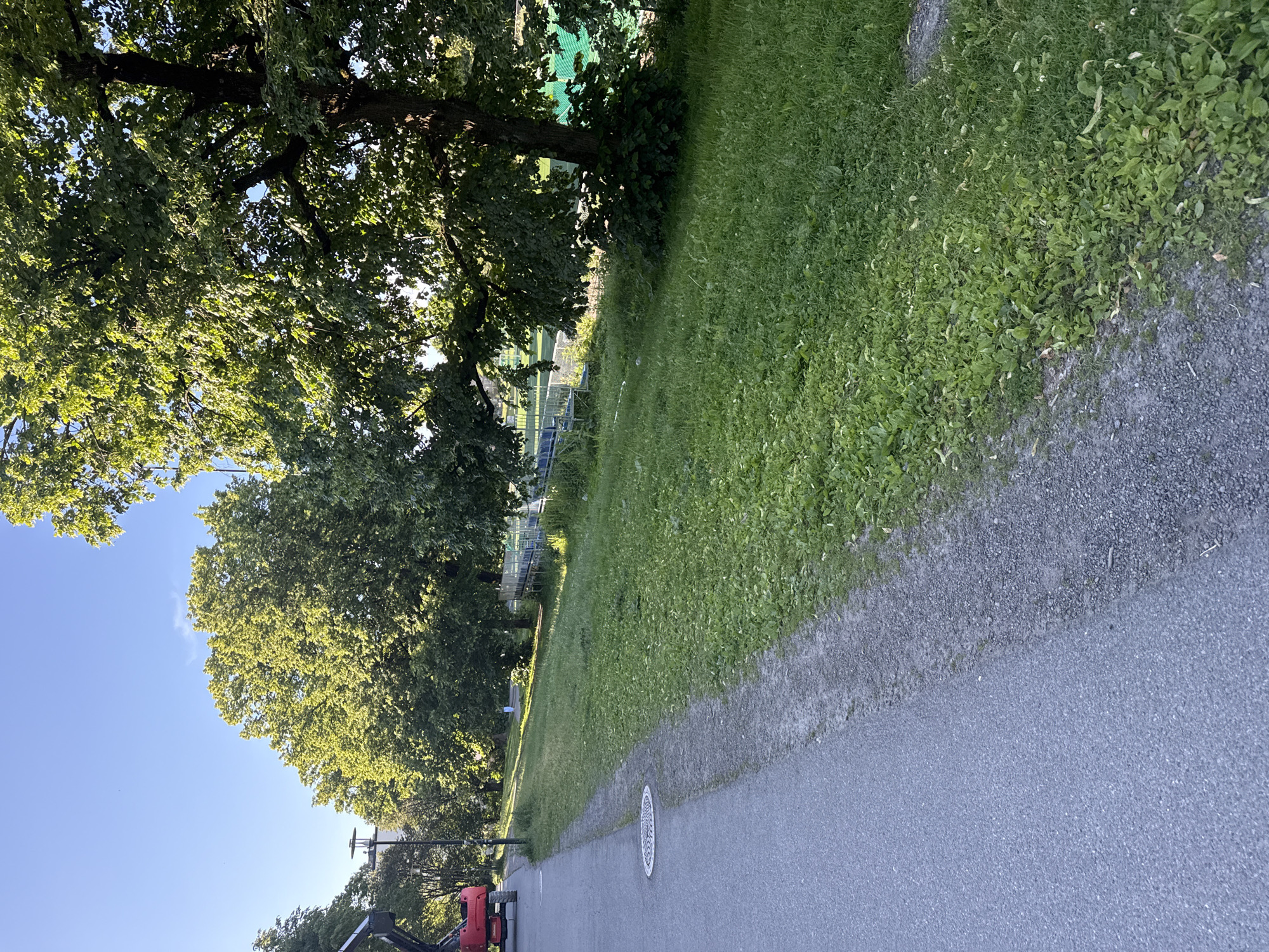 The image shows a sunny day with clear blue skies. A paved path runs along the left side of the frame, bordered by a strip of gravel and then a grassy area. Several large, mature trees with lush green foliage line the right side of the path, casting shadows on the grass. In the background, there are structures that appear to be part of a sports facility, including bleachers and fencing, with a green sports field visible beyond. A red construction vehicle is parked near the left edge of the image. The overall scene suggests a public park or recreational area within a city.