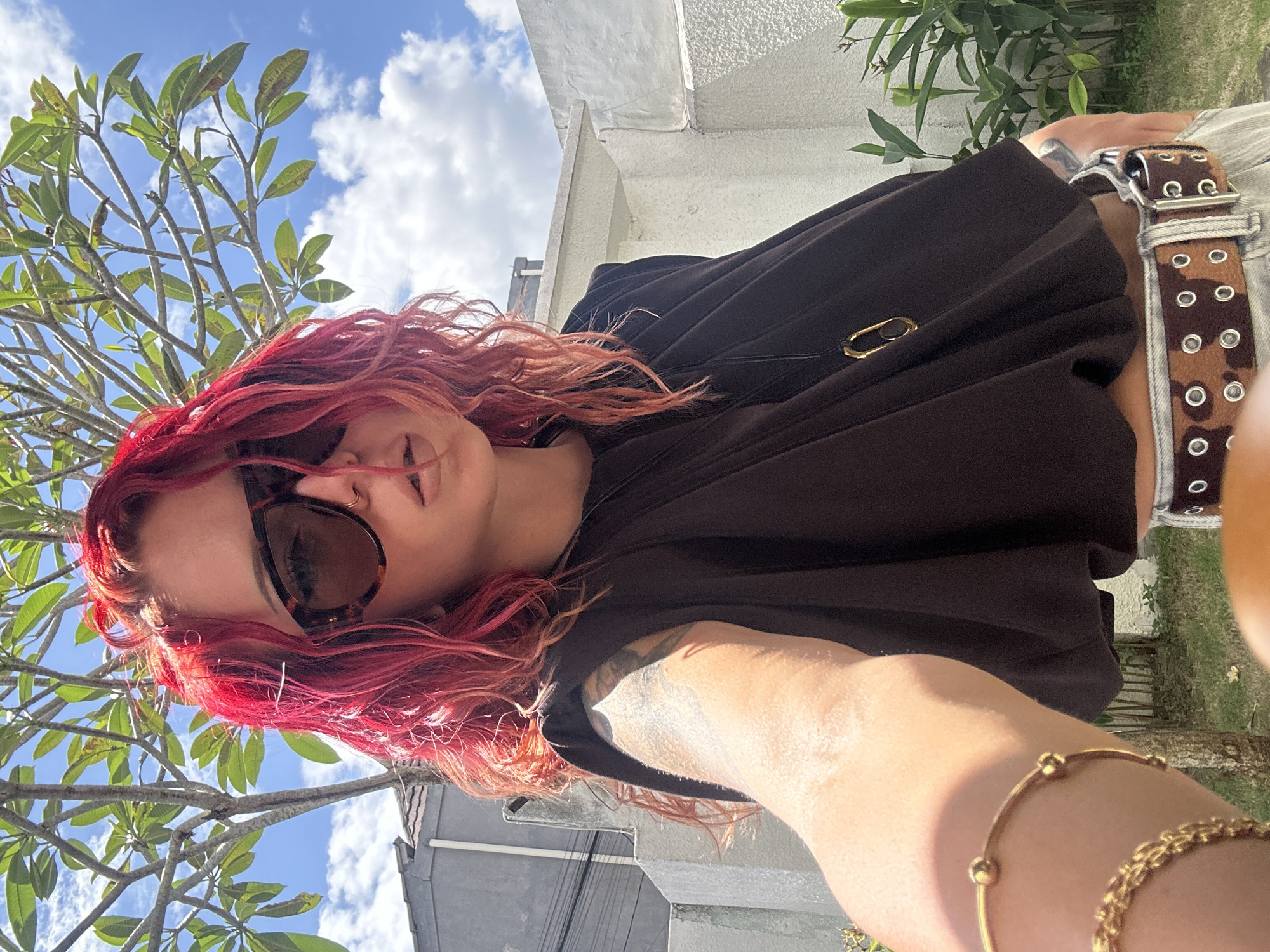 The image depicts a woman with red hair wearing sunglasses and a brown shirt. She is outdoors, with a tree visible in the background along with a building. The sky is blue with white clouds. Part of her outfit including a belt is shown. It is daytime. The location is North Kuta, Indonesia.