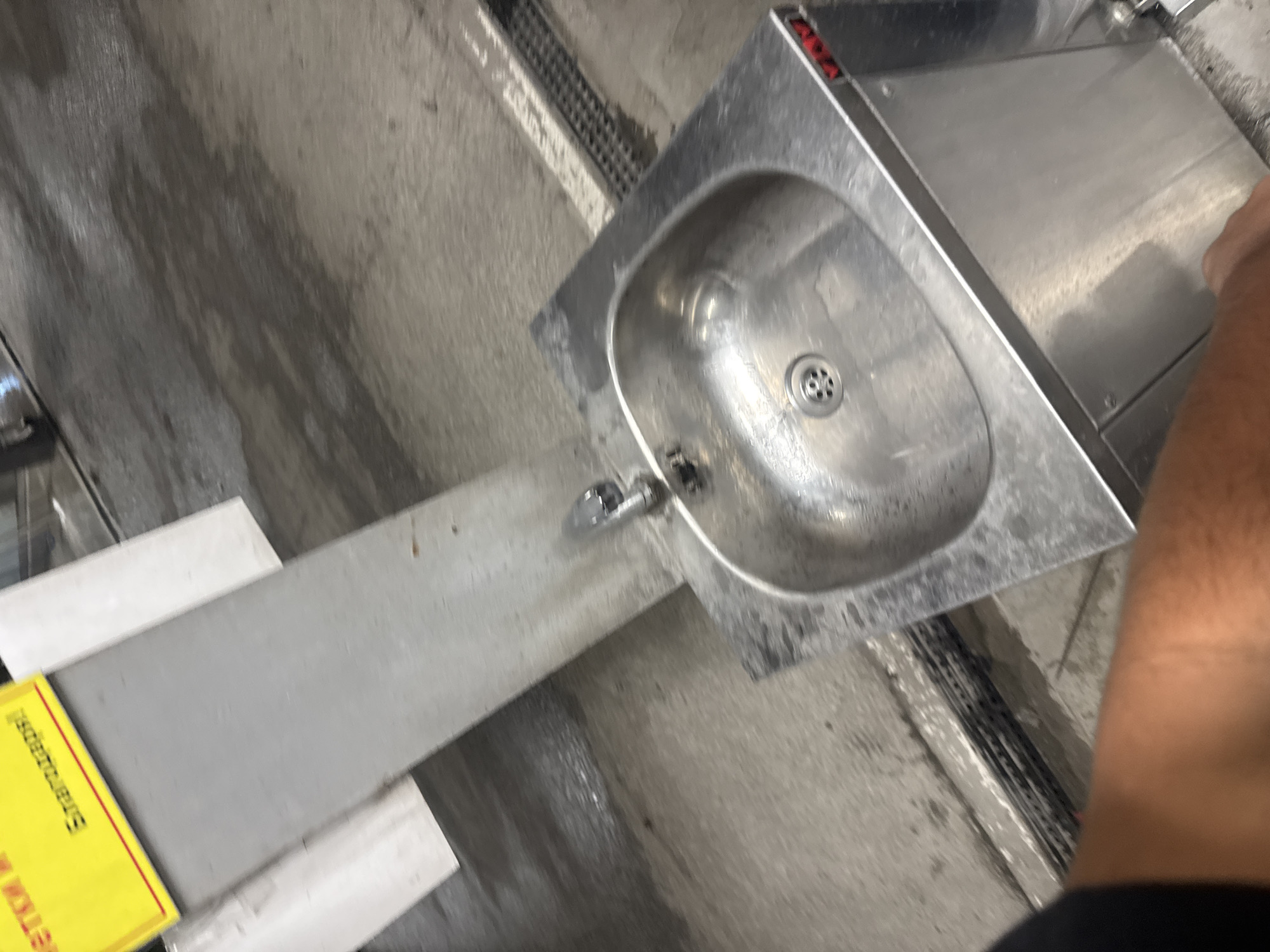 The image displays a utilitarian indoor setting, centered around a stainless steel sink unit. The sink, equipped with a drain and a single faucet, is integrated into a metal counter. The floor surrounding the sink area is concrete and appears wet, showing streaks and puddles that suggest recent cleaning or water spills. A long metal draining grate is visible on the floor, running parallel to the sink unit, designed to collect water.
On the far right, a person's bare forearm and a fragment of dark clothing are visible, indicating human presence. To the left of the sink, a yellow sign with black text is partially visible. The discernible text reads "Strahlungsge...", which is part of the German word "Strahlungsgefahren," translating to "radiation hazards." A small, blurry red label or marking is also present on the back panel of the sink unit.
The overall environment suggests a functional space, possibly a laboratory, industrial kitchen, or workshop, where hygiene and specialized safety protocols might be in place due to the presence of the warning sign. The wet floor implies ongoing or recent activities involving water. The presence of German text in Plovdiv, Bulgaria, suggests either internationally sourced equipment or a facility with specific international standards.