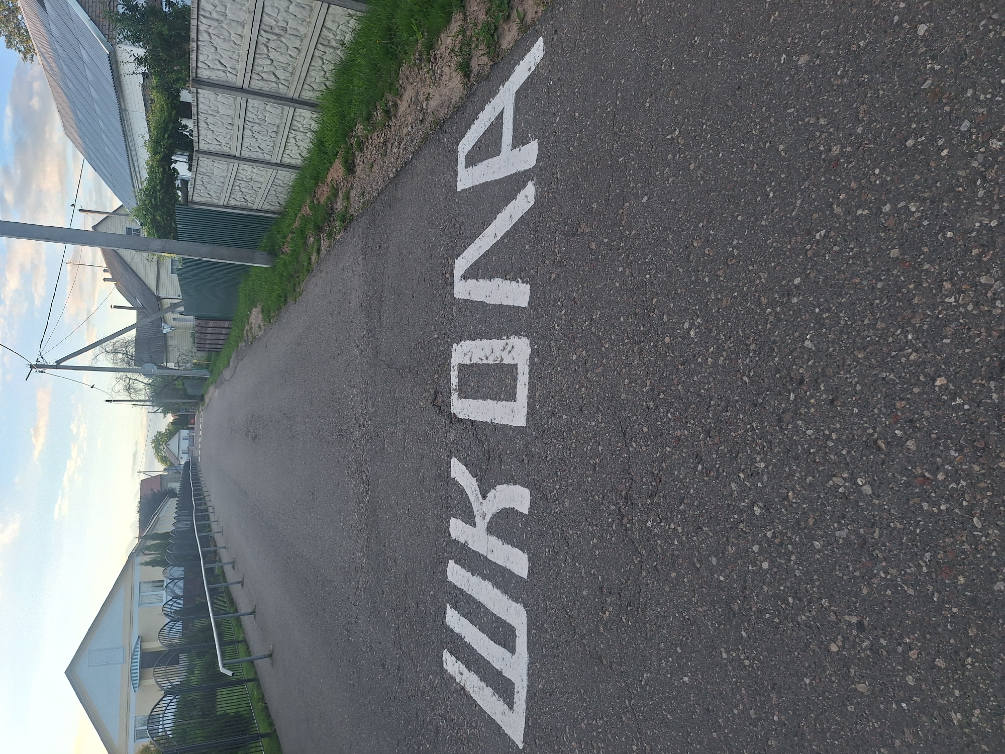 The image shows a road in what appears to be a rural or suburban area of Капыльскі сельскі Савет, Belarus. White letters painted on the asphalt road spell out "ШКОЛА," which translates to "SCHOOL" in Russian. The road is bordered by houses and fences on both sides. On the left, there is a black metal fence with a white handrail, and on the right, there is a concrete fence. The sky is visible in the background, with some clouds. The lighting suggests it is either early morning or late afternoon. There are no people or vehicles visible in the image.
