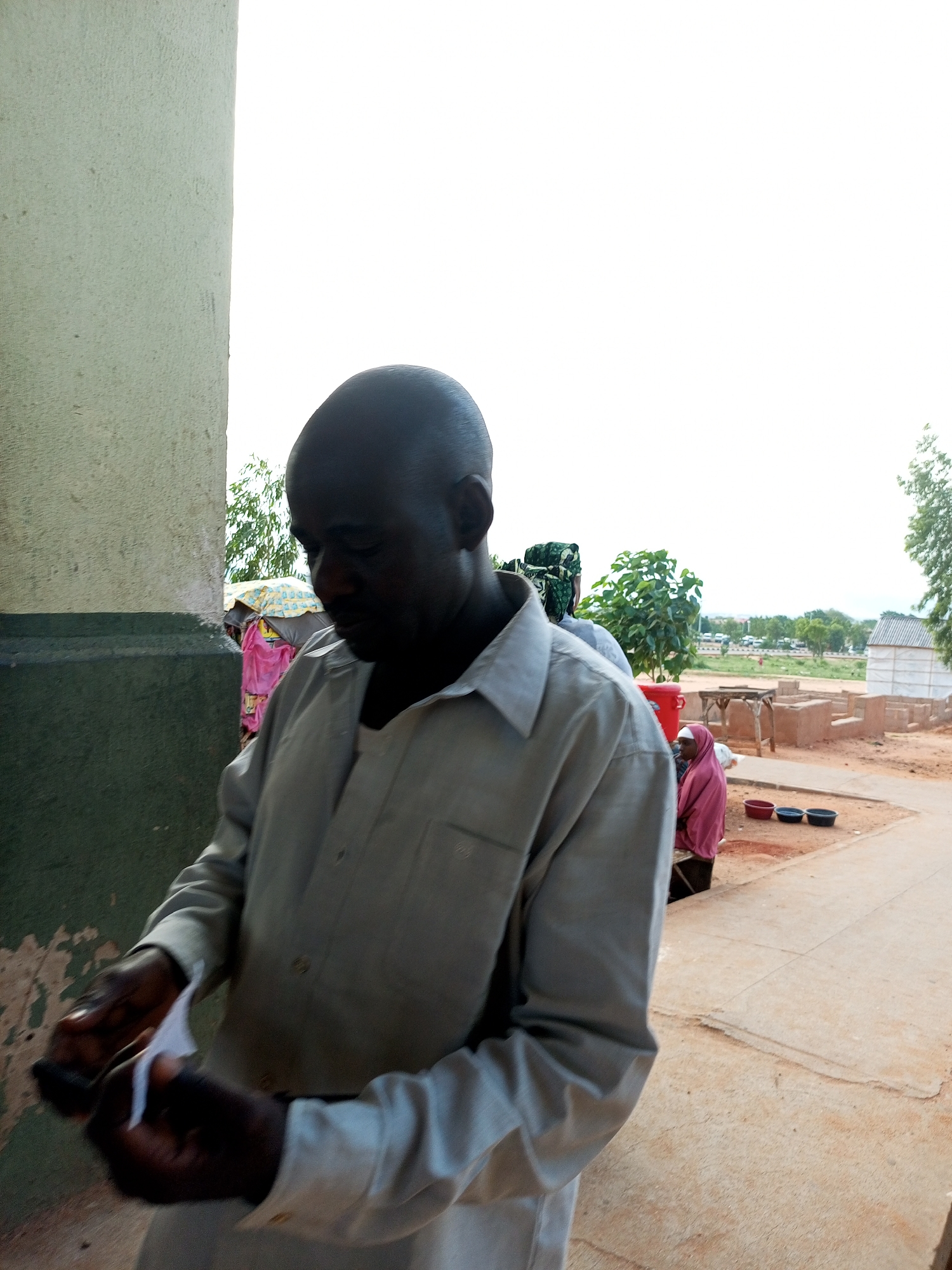This image captures an outdoor scene in Gombe, Nigeria. The primary subject is a dark-skinned man with a shaved head, wearing a light gray collared shirt. He is looking down and appears to be holding a small white object, possibly paper, in his hands, which are dark-skinned. The man is positioned in the foreground, with a textured, light-colored wall to his left and a pathway or dirt ground in front of him.
In the background, several other people are visible. To the left, behind the main subject, a person is partially obscured, wearing a pink garment and a headscarf. Further back, there are structures that appear to be small buildings or stalls, with some colorful fabrics, including pink and yellow, visible. There's also a red bucket and some dark containers on the ground. Beyond these structures, trees and a distant treeline are present, suggesting a somewhat rural or semi-urban environment. The sky is bright and appears to be daylight, likely a clear or partly cloudy day. The overall impression is that of a public space, possibly a market or a gathering area, with people engaged in daily activities. The man's posture and focus on the object in his hands suggest he might be performing a task or examining something. There is no visible text in the image.