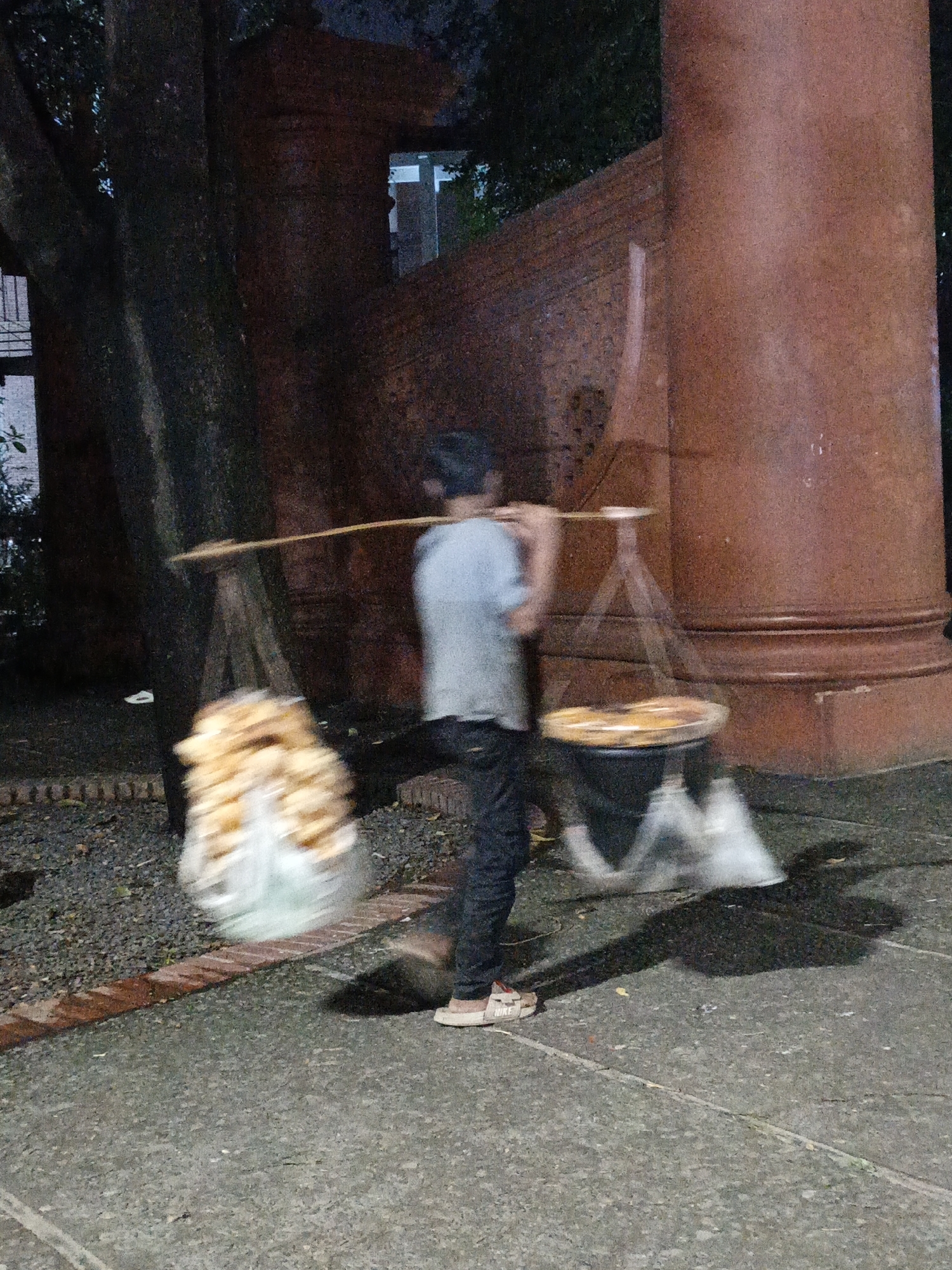 A blurry photo shows a vendor in Unknown, Unknown carrying baked goods on a shoulder pole. The vendor, wearing a light-colored shirt and dark pants, walks on a paved surface with a brick border. The vendor is carrying a large bundle of bread on one side and what appears to be round cakes or pastries on the other. A large tree and a reddish-brown cylindrical structure are in the background.