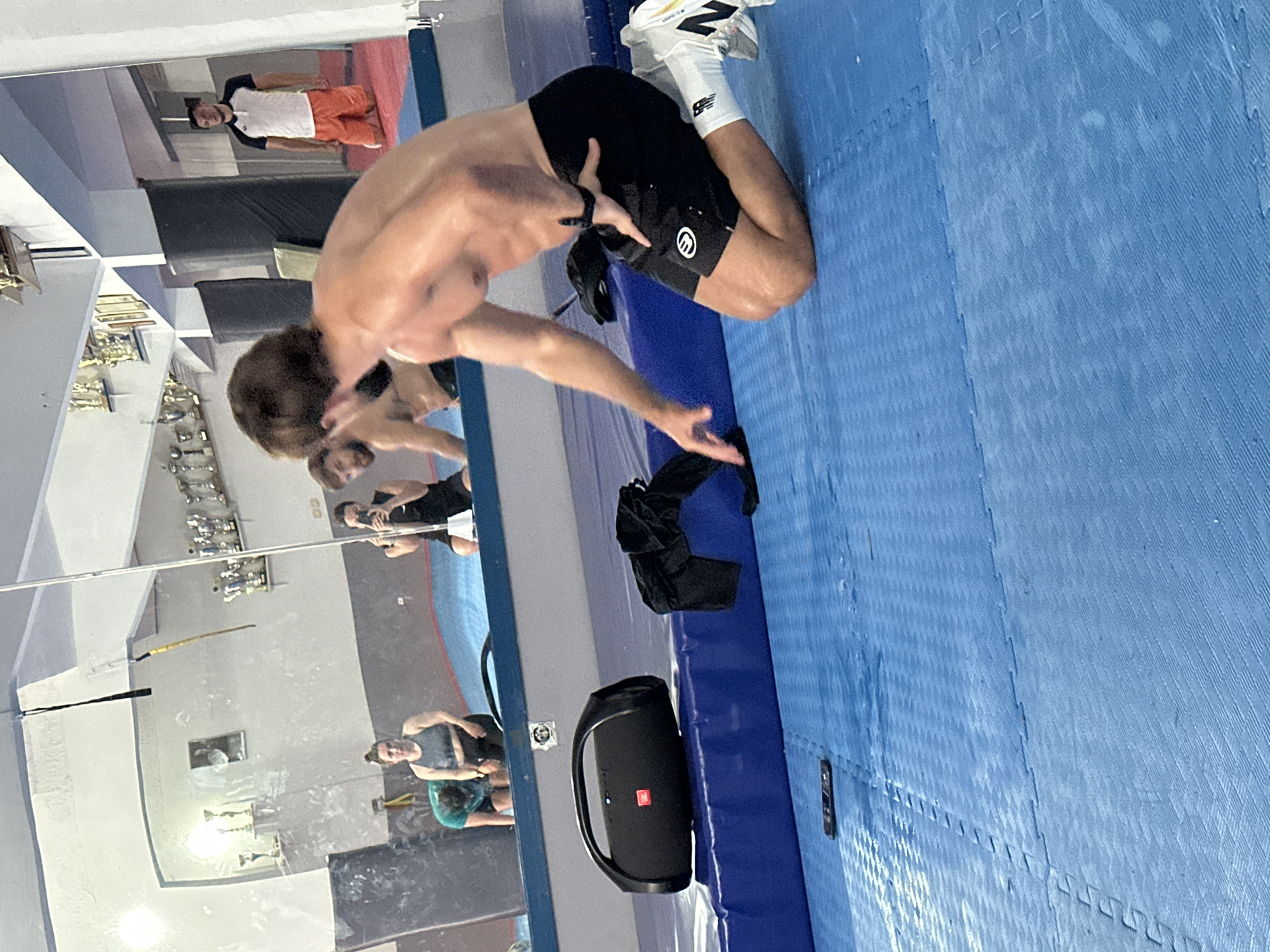 A man with brown hair and a muscular physique is shown in a dynamic pose inside what appears to be a training facility, possibly in Budva, Montenegro, given the request in the prompt. He is wearing black athletic shorts, white socks, and New Balance sneakers. He is positioned on a blue padded mat with a textured surface. A black JBL speaker and a black item of clothing (possibly a jacket) are on the floor near him. The background features a mirror that reflects a training room with other people and trophies on a shelf. One man is wearing a white t-shirt and orange shorts and standing in the reflection.