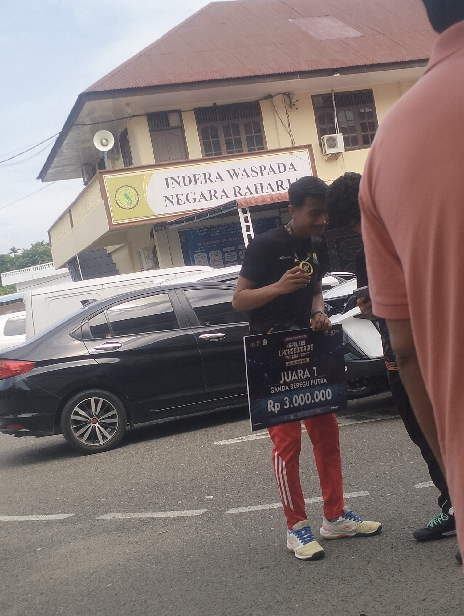 A young man, dressed in a black athletic shirt and red pants with white stripes, stands on a paved road in front of a building. He holds a sign that reads "JUARA 1 GANDA BEREGU PUTRA Rp 3.000.000" which translates to "1st Place Men's Doubles Team Rp 3,000,000." He is wearing a medal around his neck and smiles. Behind him, a black car is parked, and the building appears to be an official government building with a sign that says "INDERA WASPADA NEGARA RAHARI." The scene suggests an award ceremony following a badminton tournament, possibly the "KAPOLRES LHOKSEUMAWE CUP II" as indicated on the sign.