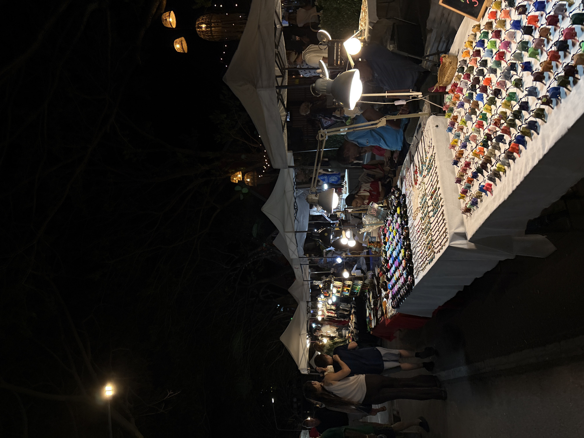 This is a nighttime shot of a market scene in Choeng Thale, Thailand. Several people are visible browsing and interacting at market stalls. The stalls are set up under white tents and are illuminated by various lights, including string lights, bright lamps, and what appear to be Edison bulbs in woven fixtures.
One stall features a large display of colorful small items, possibly sunglasses or accessories, arranged in neat rows on a white table. Another stall displays numerous necklaces and bracelets. The overall atmosphere is that of a lively night market.
The background is dark, suggesting it is late in the evening or night. Trees with dark, bare branches are visible on the left side, adding to the nighttime ambiance. The ground appears to be a paved path. Some of the people are close together, possibly a couple, in the foreground. The scene evokes a sense of evening commerce and community gathering. There is a small sign with text on it above the stall with the colorful items.