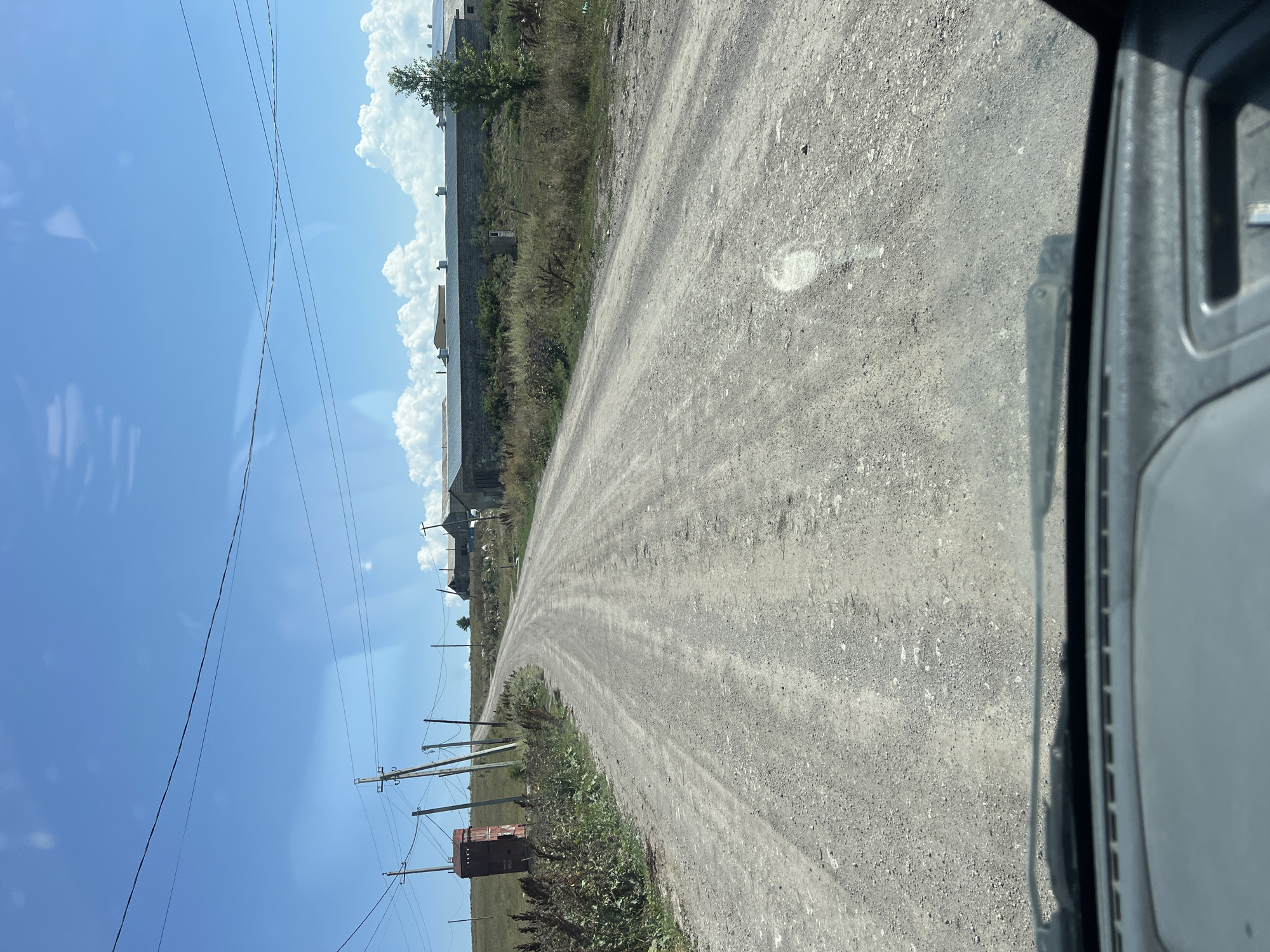 The image depicts a dirt road winding through a rural landscape under a bright blue sky with scattered white clouds. The perspective is from a vehicle, looking forward. On the left side of the road, a red brick structure, possibly a utility box or historical marker, stands next to a cluster of wooden utility poles with wires. Further down the road, a long, low-slung building with a grey, corrugated metal roof stretches across the horizon. This structure appears to be an agricultural or industrial facility, with several vents or chimneys visible on the roof. A few trees are scattered in the vicinity of the building. The surrounding terrain is characterized by dry grass and vegetation. The road itself is unpaved and appears to be in moderate condition, with visible tracks and some loose gravel. The overall impression is one of a remote, possibly agricultural setting. The location is identified as Khando, Georgia.