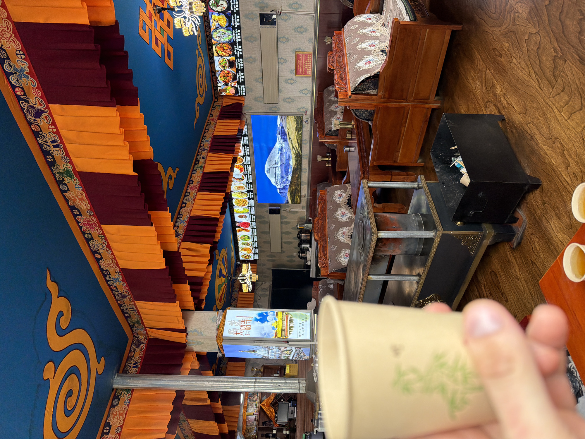 The image shows the interior of a restaurant in Jila, China. The decor includes ornate blue ceilings with yellow and maroon fabric draping, a menu displayed on a screen, and a large image of a mountain. Tables with built-in heating elements are present, along with wooden chairs and a trash receptacle. A hand holds a cup with a green design in the foreground. Two small cups containing liquid are visible on a wooden table.