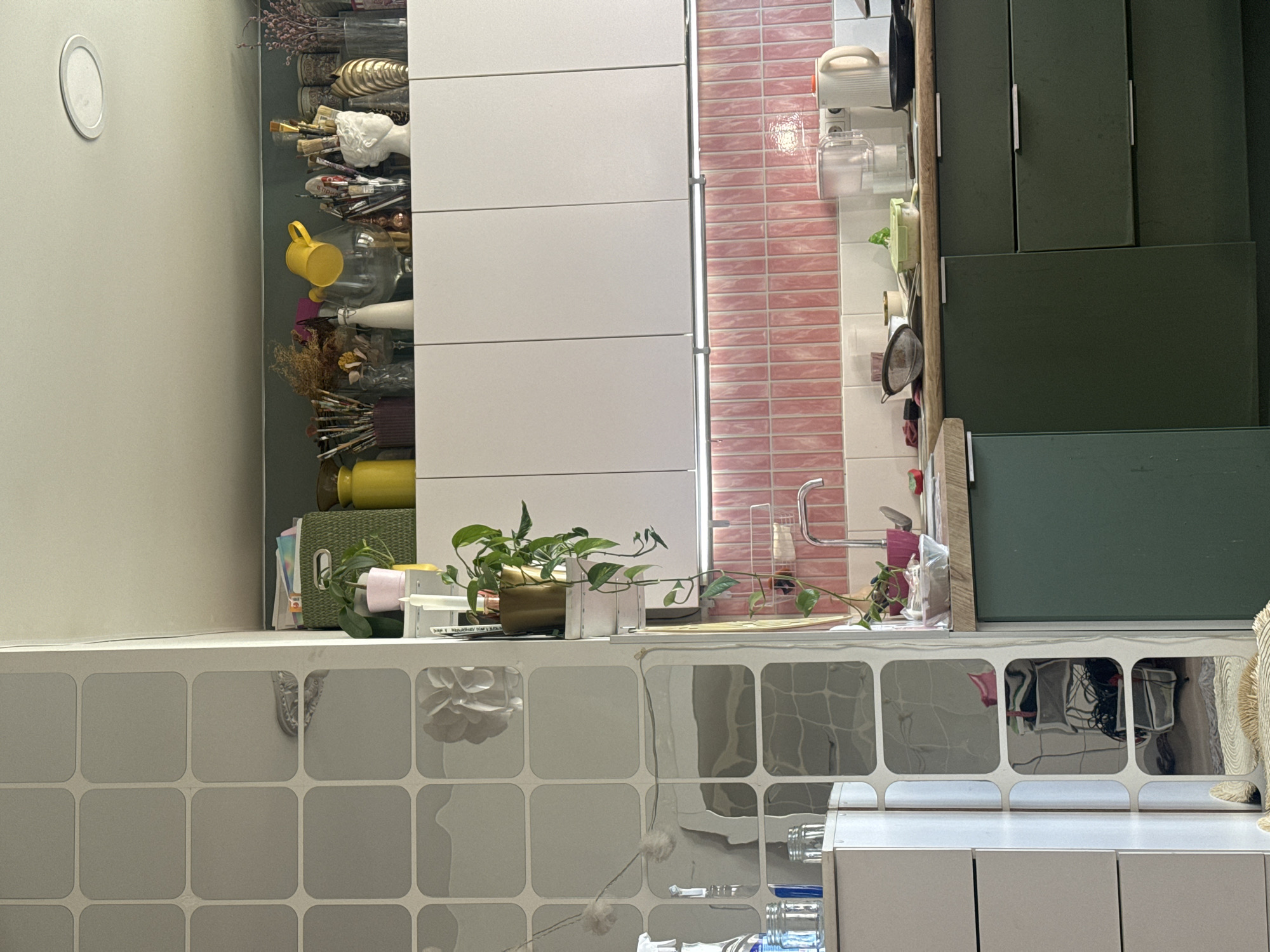 The image presents an indoor scene, likely a kitchen or utility area. On the left is a tall, narrow cabinet filled with various items, including containers, vases, and possibly art supplies. Next to it is a white, multi-drawered cabinet. Adjacent to this is a wall with pink brick-like tiles. To the right is a green cabinet. A plant is placed on the counter in front of the cabinets. The counter features a grid-like pattern with reflective surfaces that mirror objects below. The location and city are not discernible from the visual data.