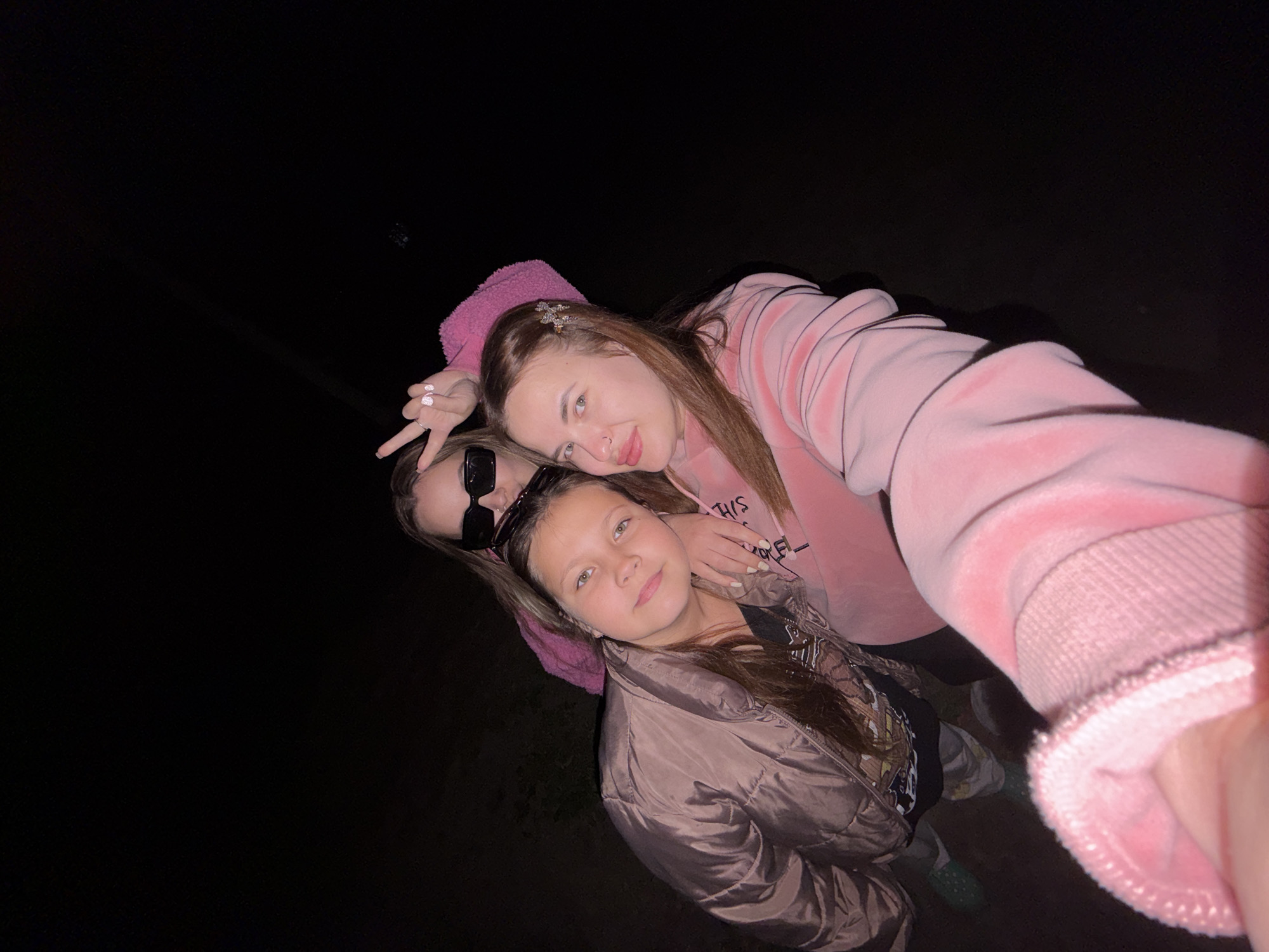 Three females are captured in a selfie-style photograph. The background is dark, possibly nighttime. The subject taking the photo is wearing a pink hooded sweatshirt. Another female in sunglasses makes a peace sign. The third female wears a light brown puffer jacket. The photograph was taken in Novopokrovskaya, Russia.