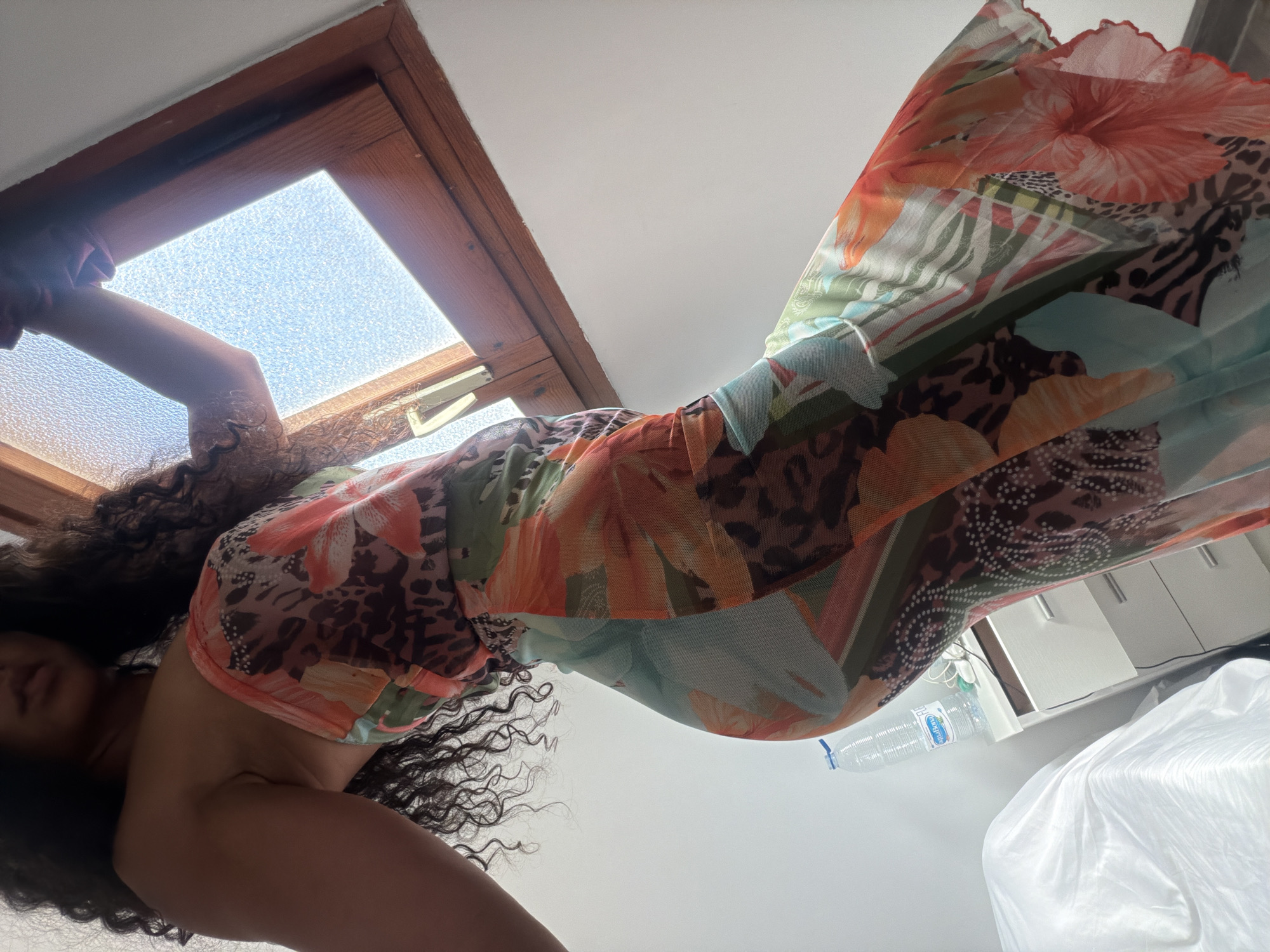 A woman with curly dark hair is wearing a colorful, patterned dress. She is standing in a room with a wooden-framed window visible in the top left corner, letting in daylight. A water bottle and some furniture are partially visible in the background. The exact location in Barcelona, Spain cannot be determined from this image.