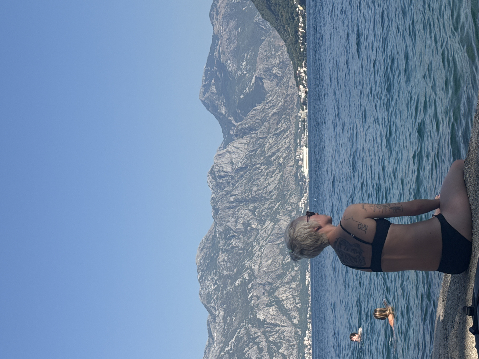 A woman with blonde hair pulled into a bun and wearing sunglasses and a black bikini is seated on a stone embankment, facing away from the camera. She has tattoos on her back and arms. In the blue water, two other individuals are swimming. Beyond the water, a white cruise ship is visible in the distance, near a town nestled at the base of a large, rocky mountain range. The sky is clear and blue. The scene appears to be a coastal area in or near Perast, Montenegro.
