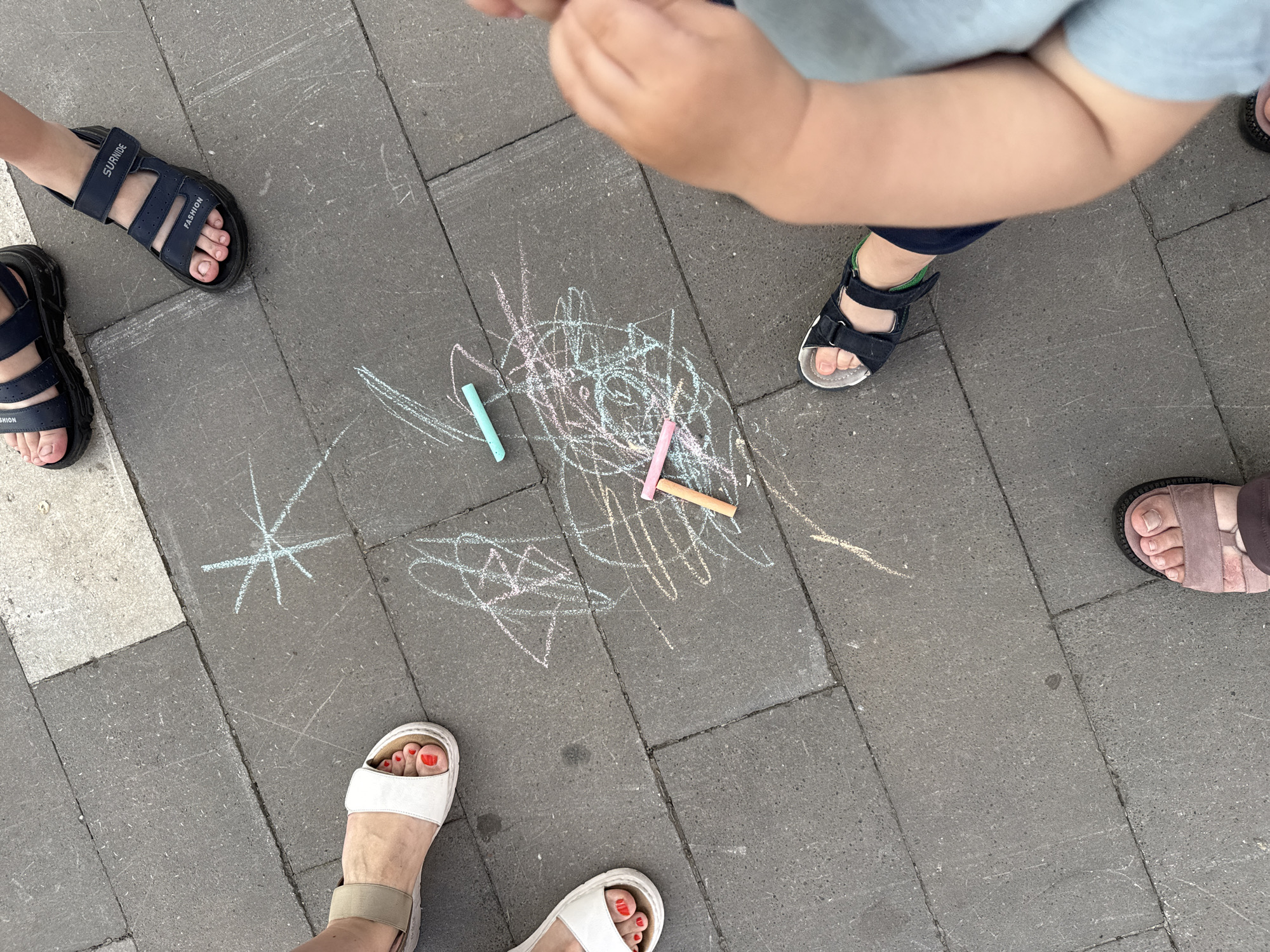 The image shows a top-down view of several people's feet standing on a paved surface, likely a sidewalk or walkway. The paving stones are dark gray and rectangular, laid in a staggered pattern. Chalk drawings, including a star-like shape and scribbled lines in various colors (white, pink, light blue, and yellow), are visible on the stones. Two pieces of chalk, one pink and one light blue, are lying on the ground amidst the drawings. Several pairs of feet are visible, wearing sandals. One person is wearing white sandals with red-painted toenails. Another person is wearing dark blue sandals with Velcro straps, and the word "FASHION" is visible on one strap. A third pair of feet is wearing dark blue sandals, and the word "SURNIDE" is visible on one strap. A fourth pair of feet is wearing a pinkish-brown sandal. The lower legs and arms of a small child are also visible in the frame. The scene suggests an outdoor activity, possibly a public space in Krasnoyarsk, Russia, where children have been drawing with chalk.