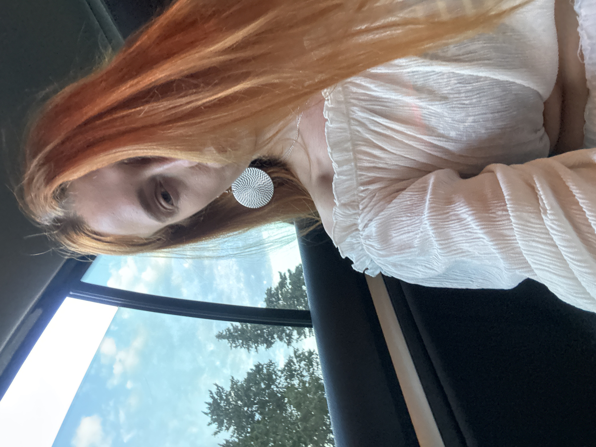 A young adult female with long, bright orange hair, partially obscuring the right side of her face, is depicted in a close-up selfie taken from inside a vehicle. Her visible left eye is directed towards the camera. She wears a white, off-the-shoulder top with a textured, crinkled fabric and a ruffled elastic neckline. A large, circular silver-toned earring with an embossed, patterned surface hangs from her left ear, and a thin silver chain necklace is visible around her neck. The background shows the interior of a car, specifically a dark-colored door panel with a thin, light-colored horizontal trim piece. Through the car window, a clear light blue sky with scattered white clouds is visible above dark green evergreen trees. The scene is illuminated by natural light, suggesting daytime. This image was taken in Petrovac centar, Montenegro.